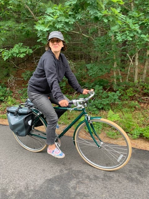 Person wearing a cap and dark clothes rides a green bicycle on a paved path through a wooded area. They have a bag attached to the bikes frame and are surrounded by greenery.