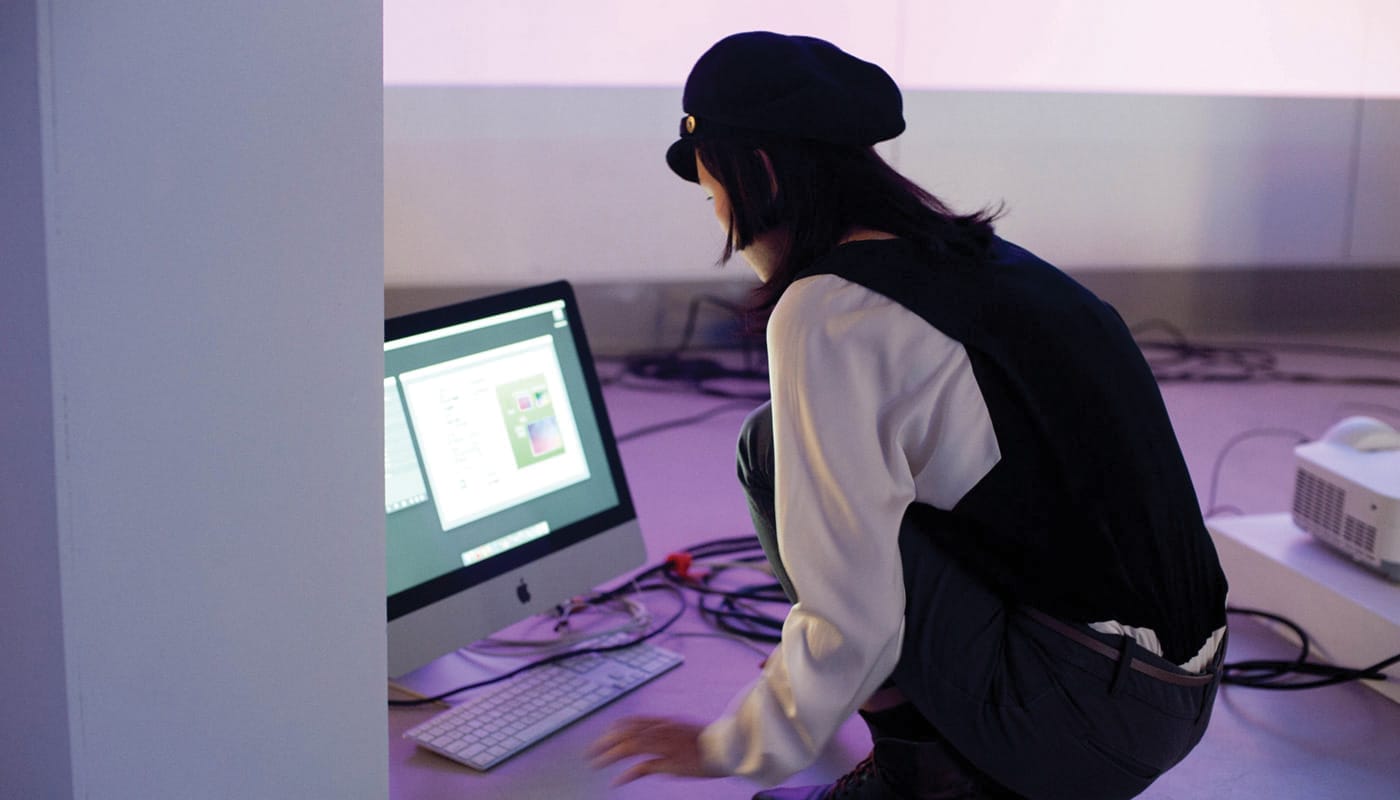 A student squats in front of a computer in a purple-lit room.