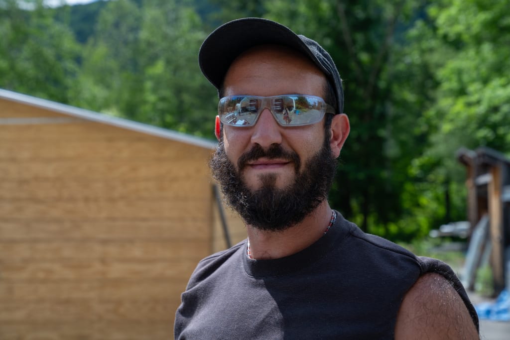 A bearded man wearing a cap and reflective sunglasses stands outdoors in front of a wooden structure, with trees and hills in the background. Hes wearing a sleeveless black shirt and smiling slightly.