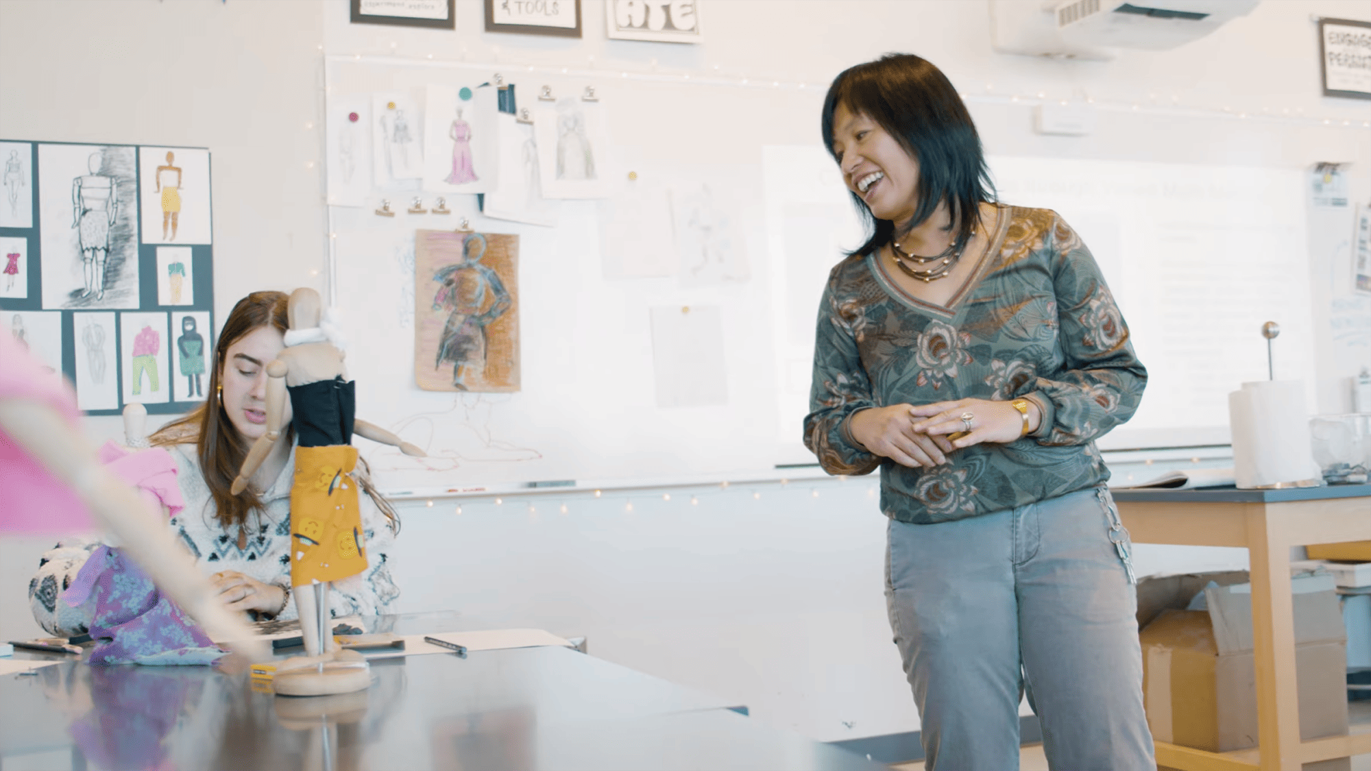 A teacher, possibly May Chau, smiles as she watches a student work on a small mannequin in yellow fabric, reflecting Art Education excellence in a classroom adorned with sketches—an inspiring scene worthy of the Frances Euphemia Thompson Award.