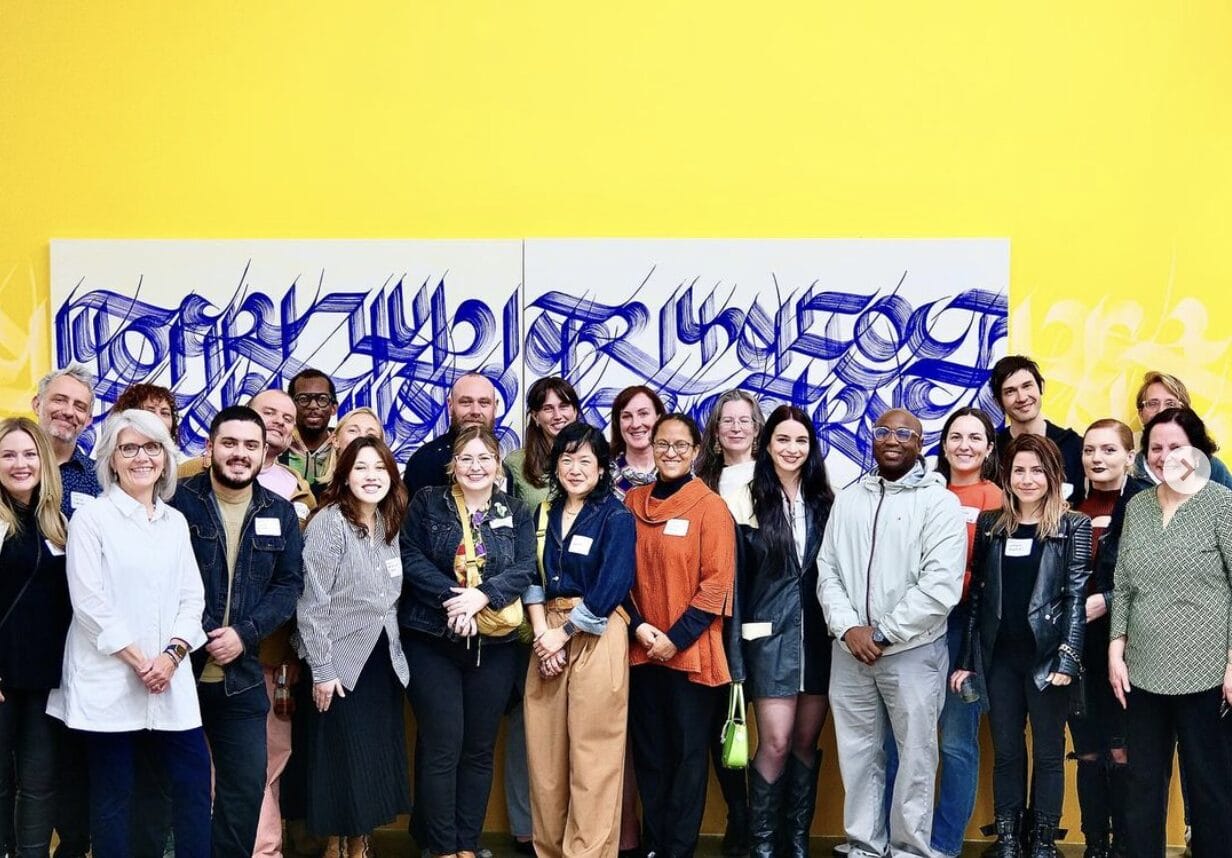 Alumni meetup group photo in a gallery setting in front of a yellow wall a large artwork.