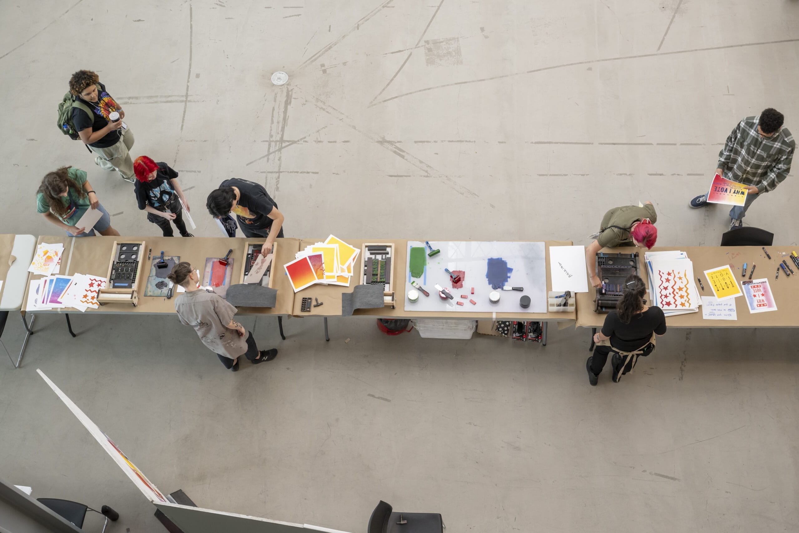 The aerial view reveals a dynamic event in a spacious indoor setting, where tables are lined with colorful papers, art supplies, and printing equipment. Individuals engage in inspecting prints or creating materials, showcasing the vibrancy of this creative space.