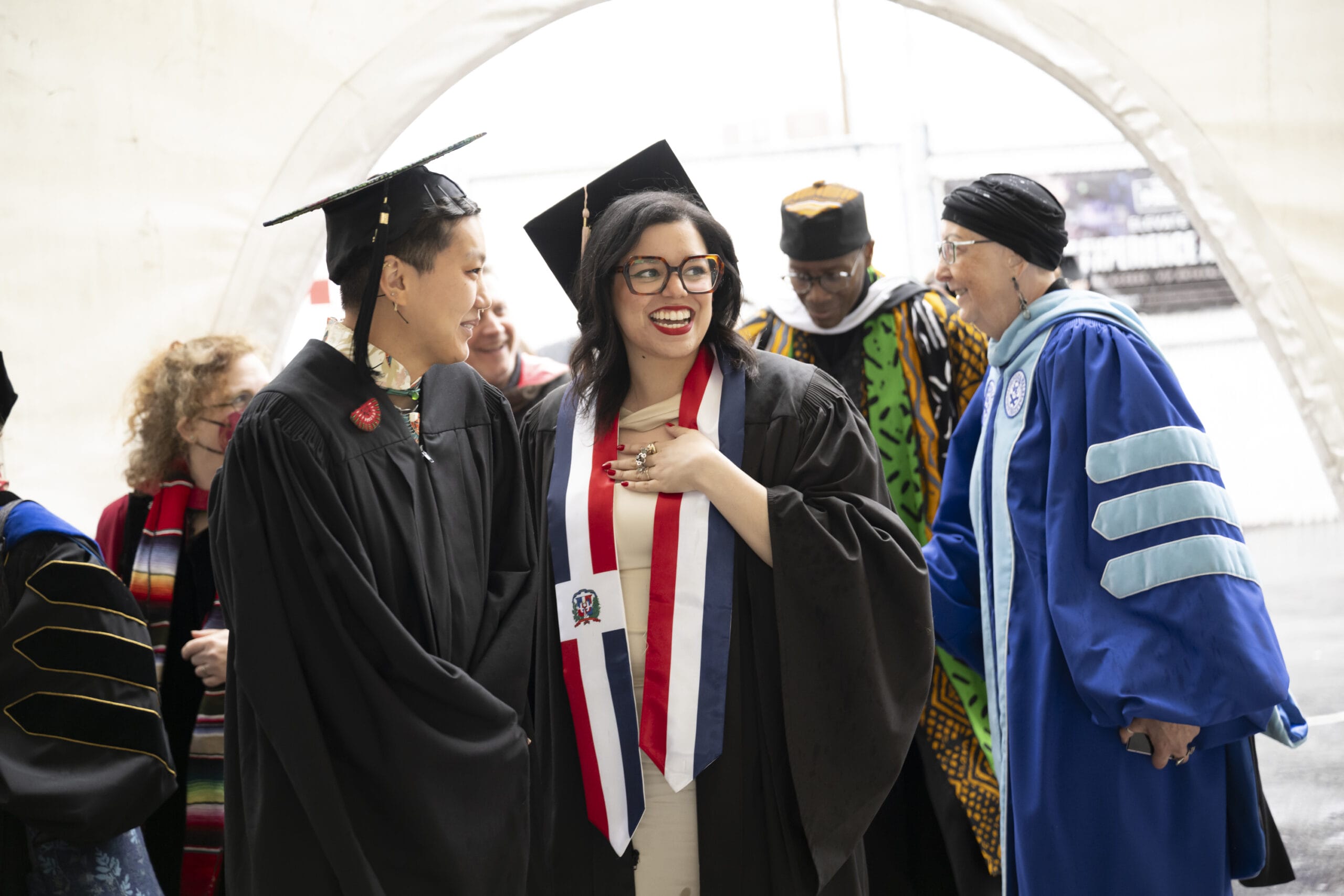 A joyous commencement unfolds as a group of graduates celebrate their achievements. Two beaming graduates in caps and gowns stand at the center, sharing a moment. Surrounding them are faculty members in academic regalia, set against a bright, blurred outdoor backdrop.