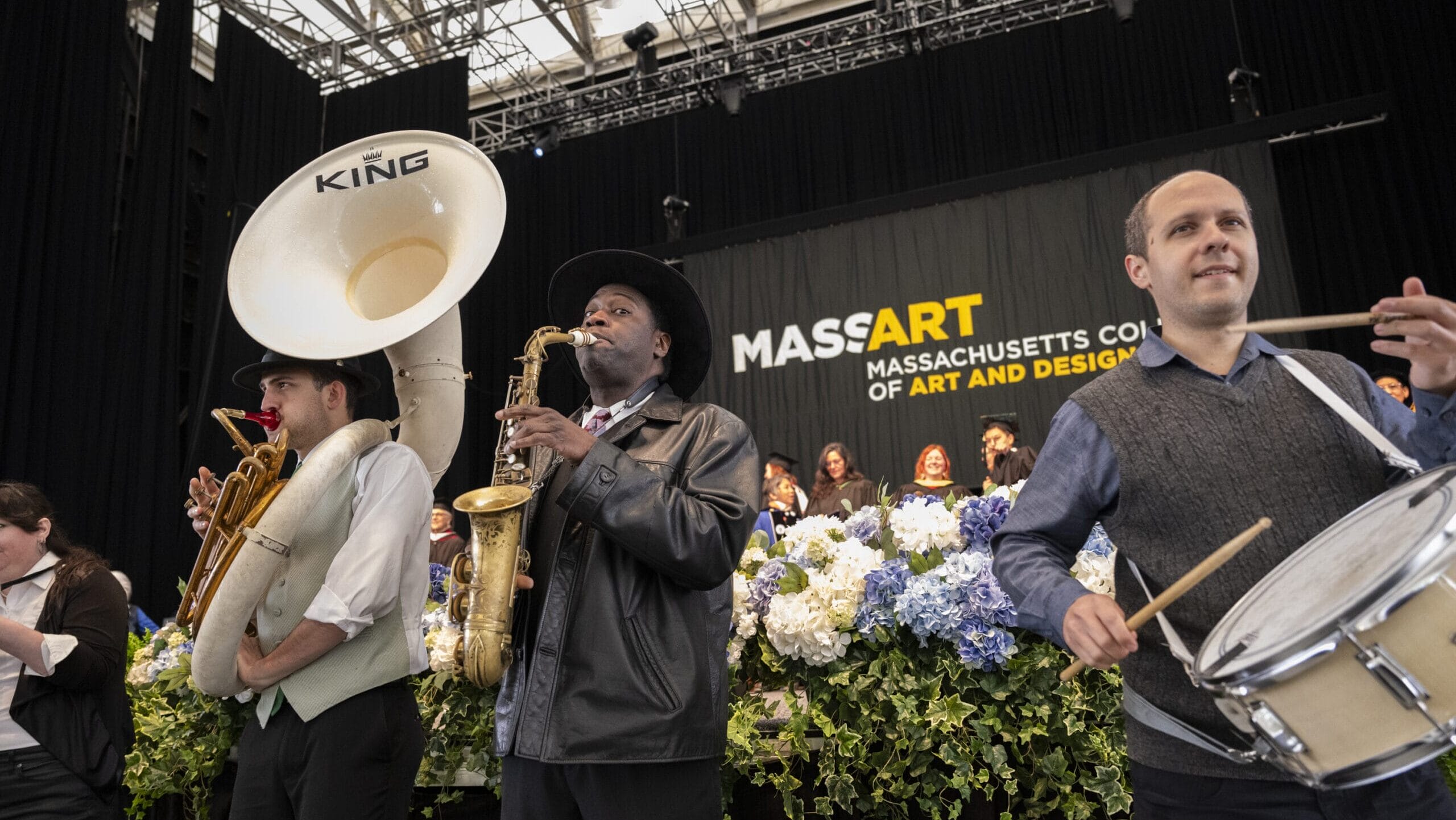 A tuba, saxophone and drum player stand in front of the Commencement stage playing music.