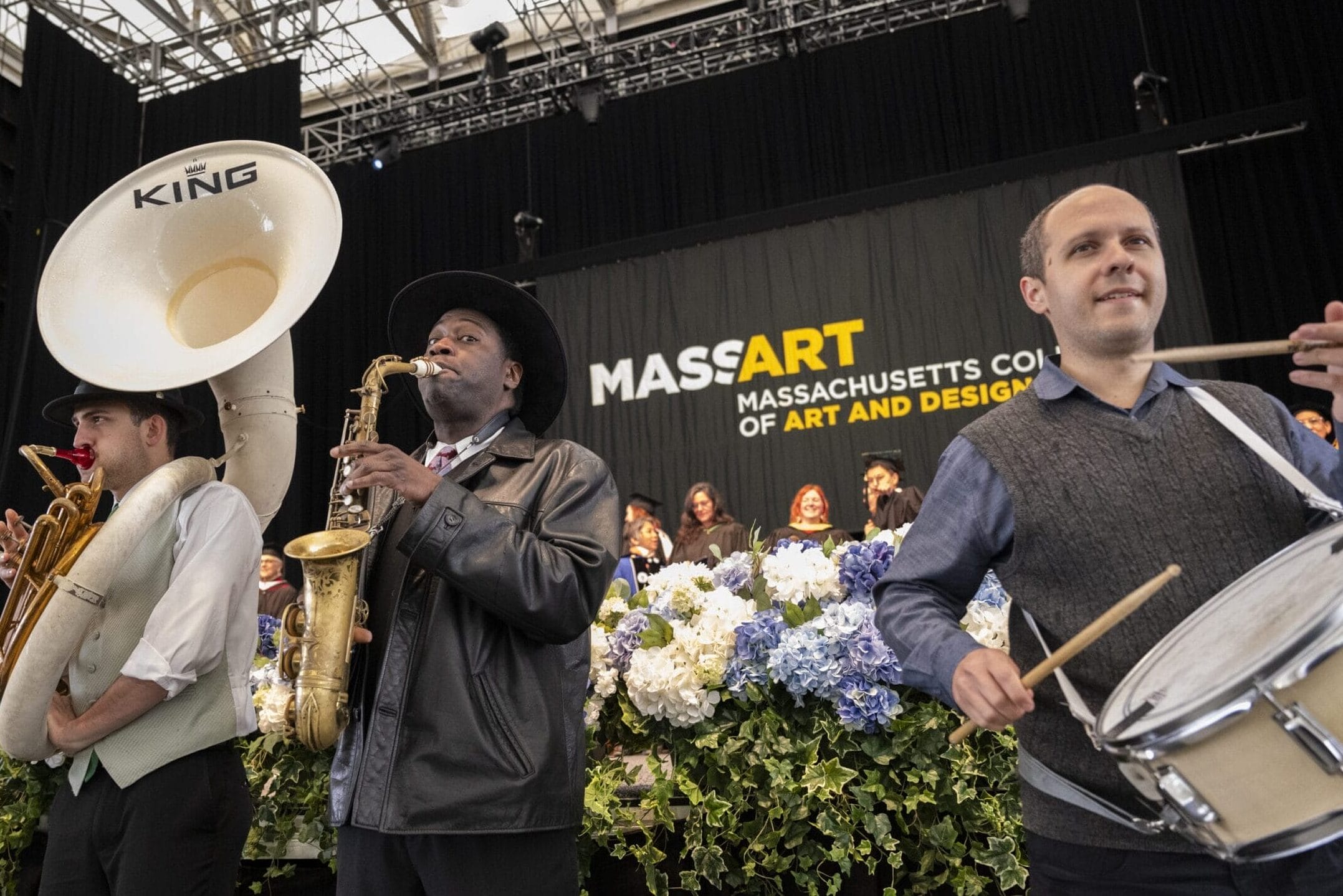 A tuba, saxophone and drum player stand in front of the Commencement stage playing music.