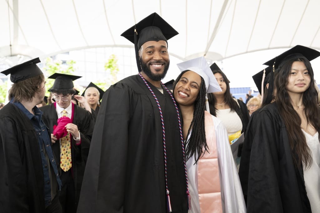 Amidst the lively graduation ceremony, a joyful moment unfolds with a man and woman in caps and gowns beaming brightly. They stand closely together among fellow graduates, capturing the airy excitement of commencement.