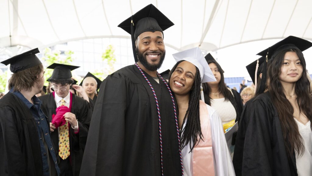 Amidst the lively graduation ceremony, a joyful moment unfolds with a man and woman in caps and gowns beaming brightly. They stand closely together among fellow graduates, capturing the airy excitement of commencement.