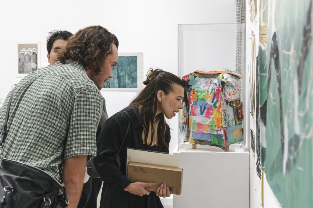 Three people observe colorful artwork on a pedestal in a gallery, immersed in a reflection of vibrant, abstract designs. One person holds a folder, leaning in to view closely, while others look on from behind. Various art pieces are displayed, enriching the creative spaces around them.