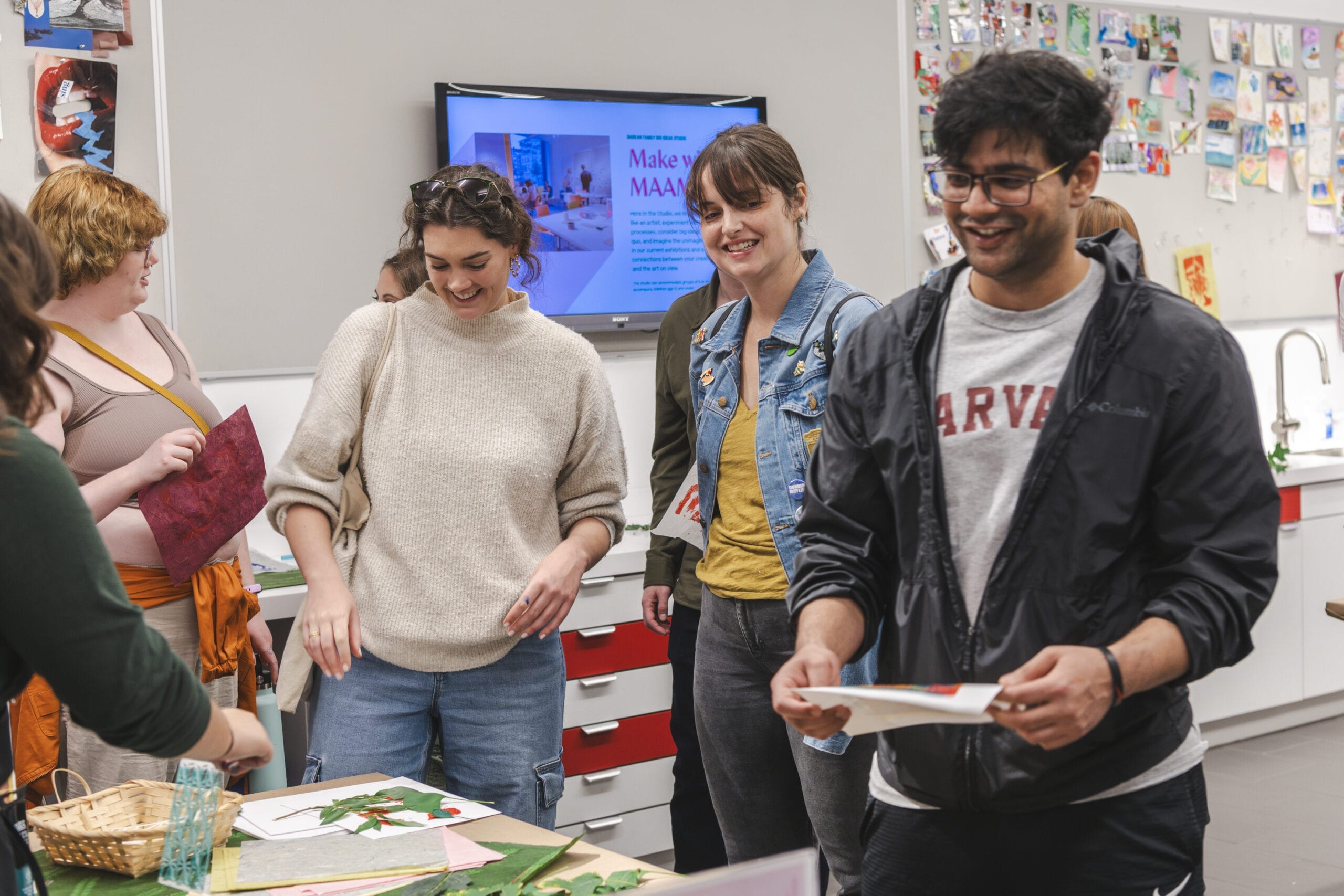 A group of people in casual clothing are gathered around a table in a creative workshop, smiling and interacting. There are art supplies on the table and colorful artwork on the walls. A monitor displays a slide titled Make Way.