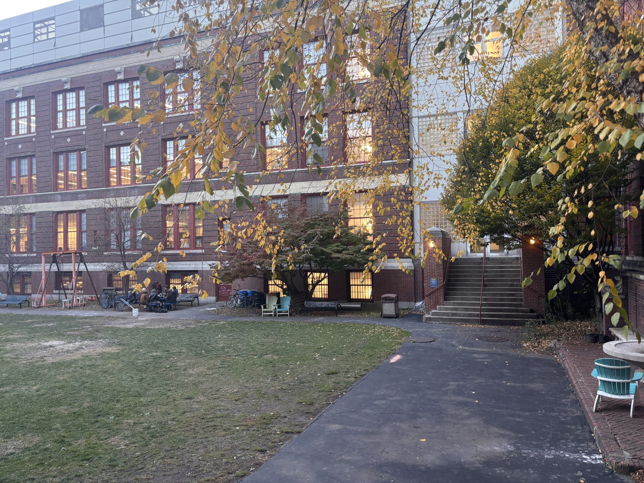 A school building with warm lighting through windows. In the foreground, a swing set, a bench, and a bike rack are visible on a grassy area. Leaves of a tree hang overhead, and a set of stairs leads to the buildings entrance on the right.