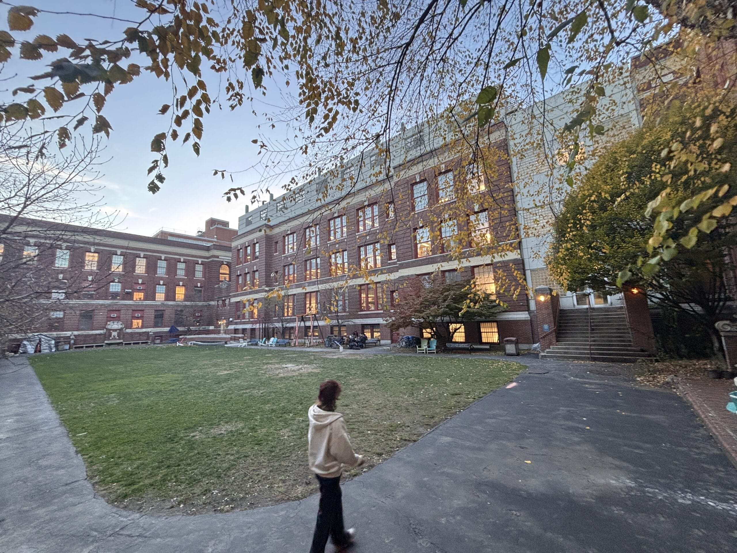 A person in a beige hoodie walks along a path in front of a large brick building at dusk. The building has multiple windows with some lights on, and a grassy area with trees surrounds the path. Leaves are falling and the sky is a dusky blue.