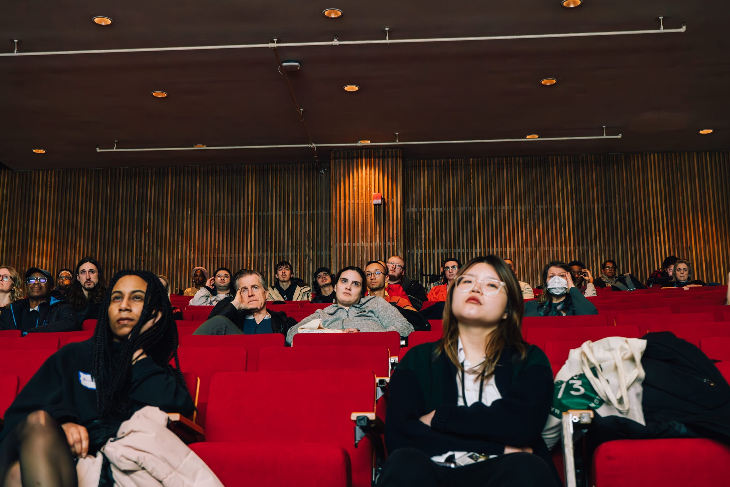 A diverse group of people sits in red theater seats, attentively enjoying the event. Some wear lanyards, while one person dons a face mask. The space is accentuated by wooden paneling and ceiling lights, creating an inviting atmosphere.