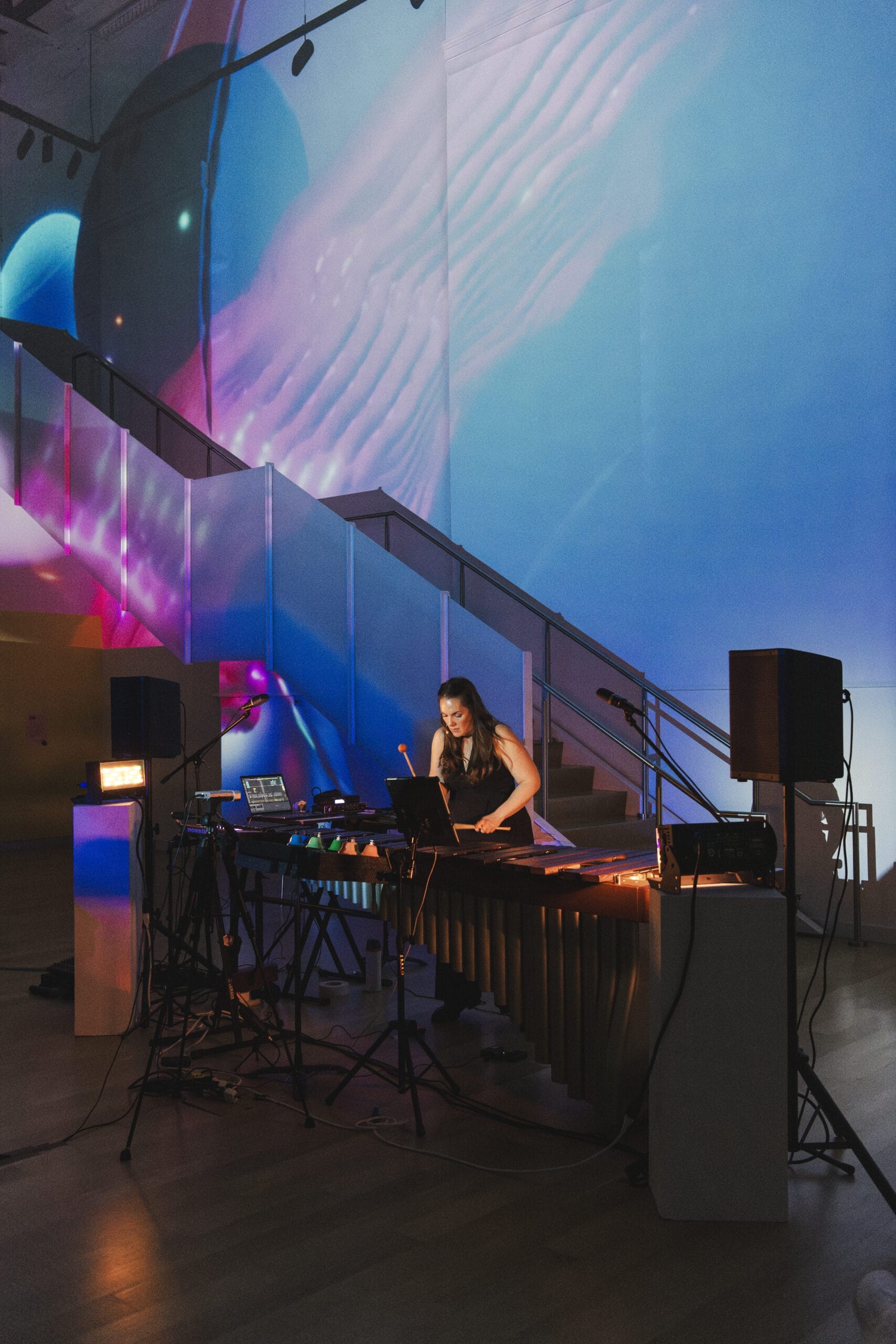 A person plays an electronic music setup, featuring a xylophone-like instrument, under colorful, abstract lighting at the base of a staircase. Surrounding them are several laptops and speakers. The atmosphere of the event is vibrant and artsy, turning the space into a creative haven.