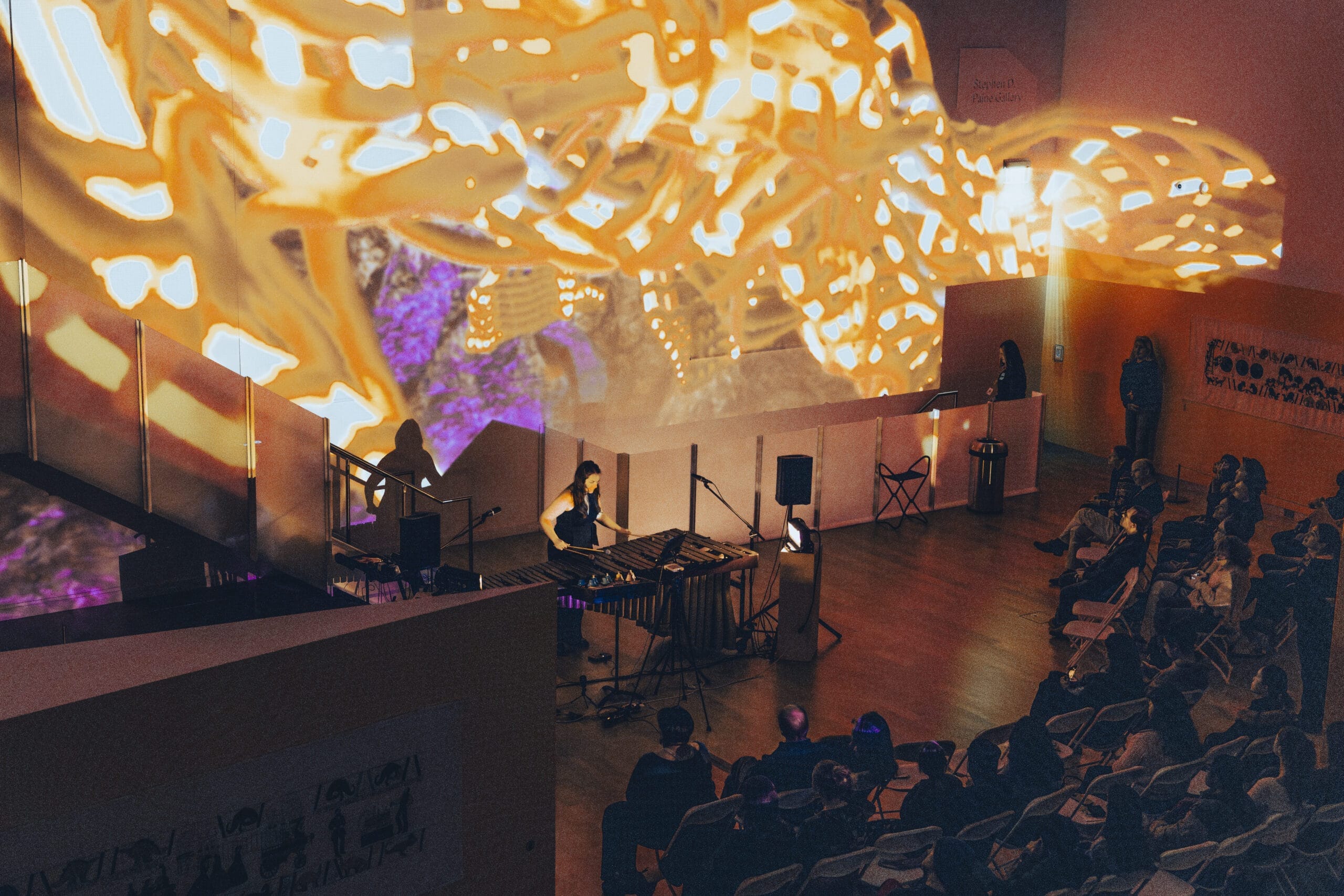 A performer plays a marimba on stage, surrounded by a seated audience. The space features vibrant, abstract projections in warm yellow tones on the walls, creating an immersive and artistic atmosphere for the event.