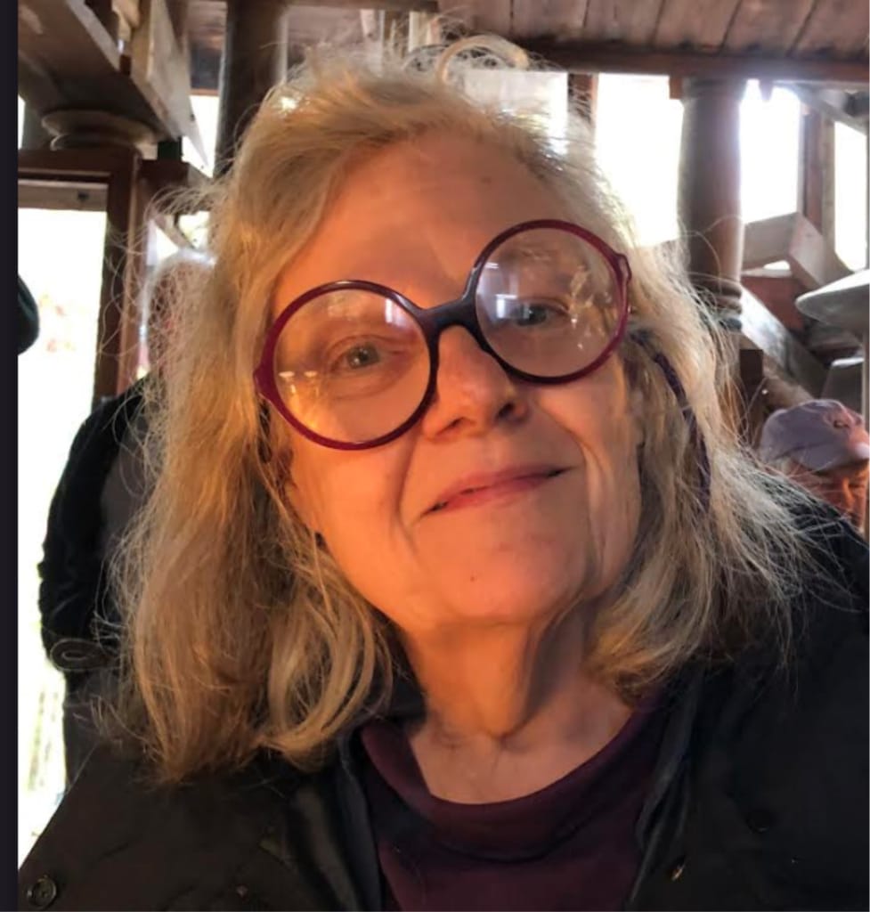 A woman with long, light hair and large round glasses smiles softly at the camera. She is indoors, wearing a dark top, and the background features wooden beams with a warm, soft light.