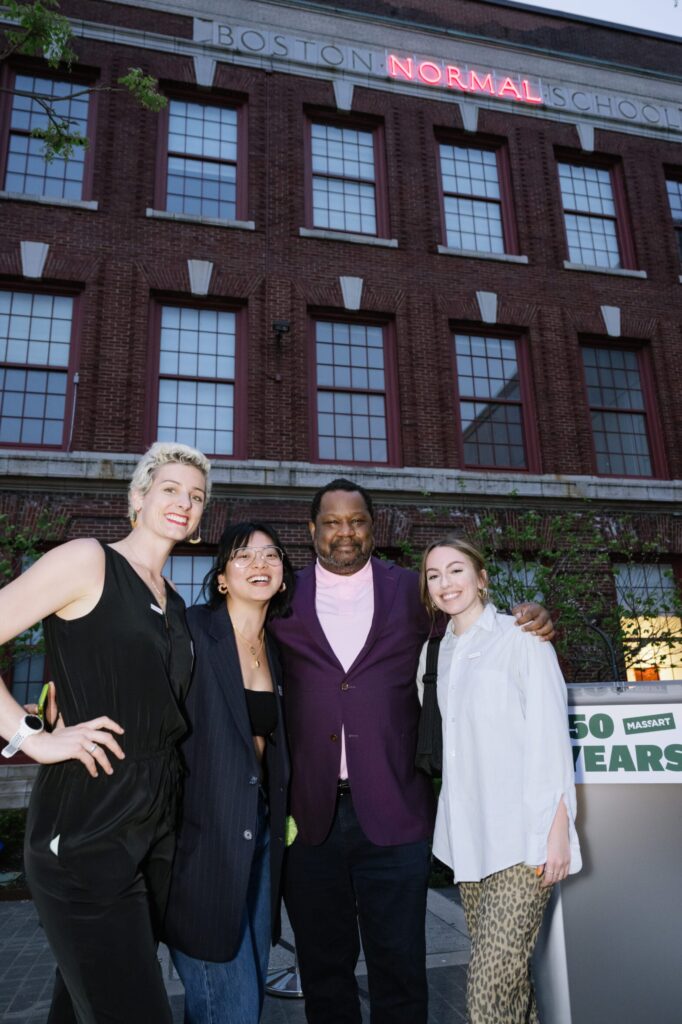Four people are smiling and posing in front of a historic brick building with Boston Normal School written on it. A sign displaying 50 Years is in the foreground.