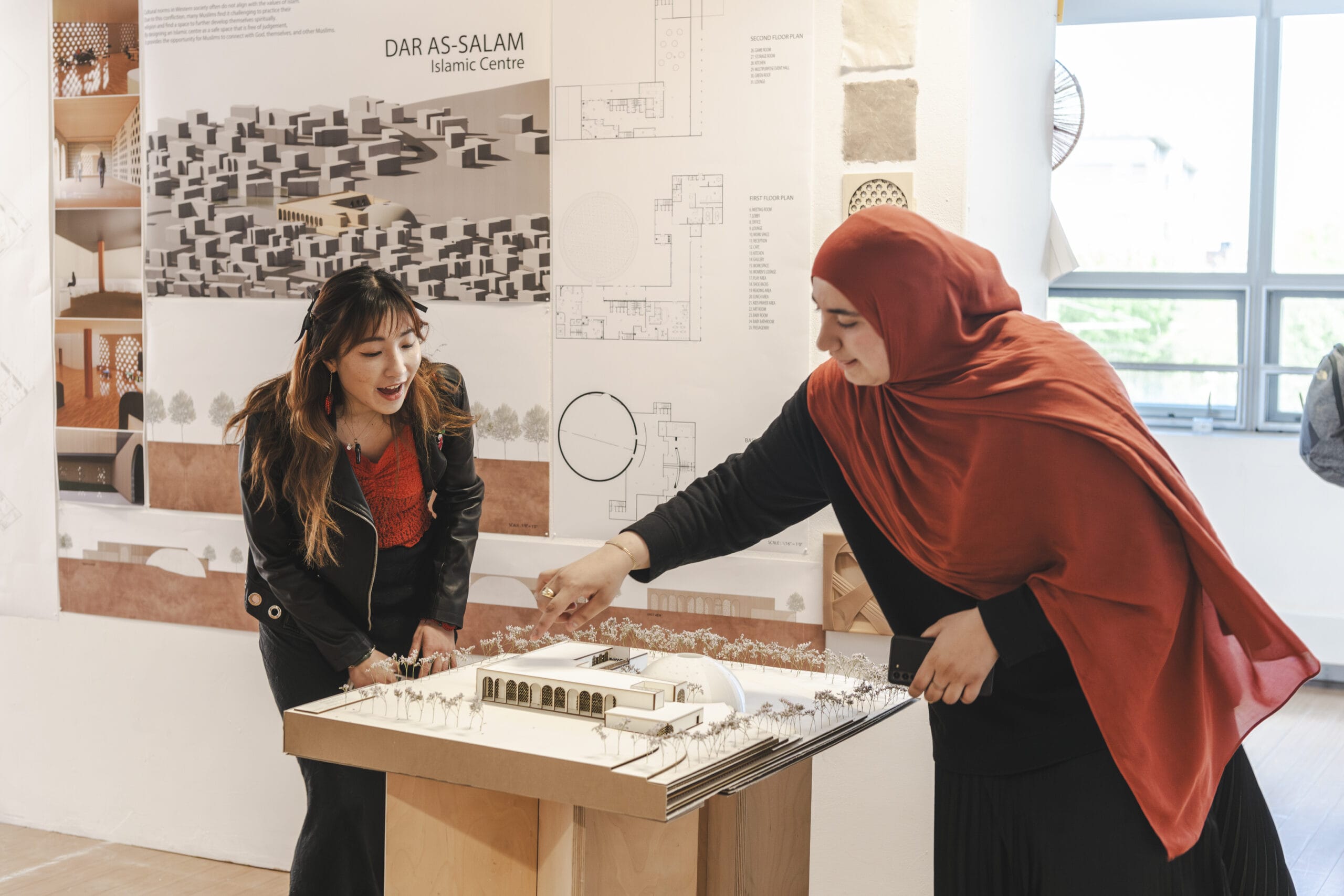 Two women examine an architectural model on a table in a bright room. The woman on the left leans in with a smile, while the woman on the right points at the model. Posters with architectural designs are displayed in the background.