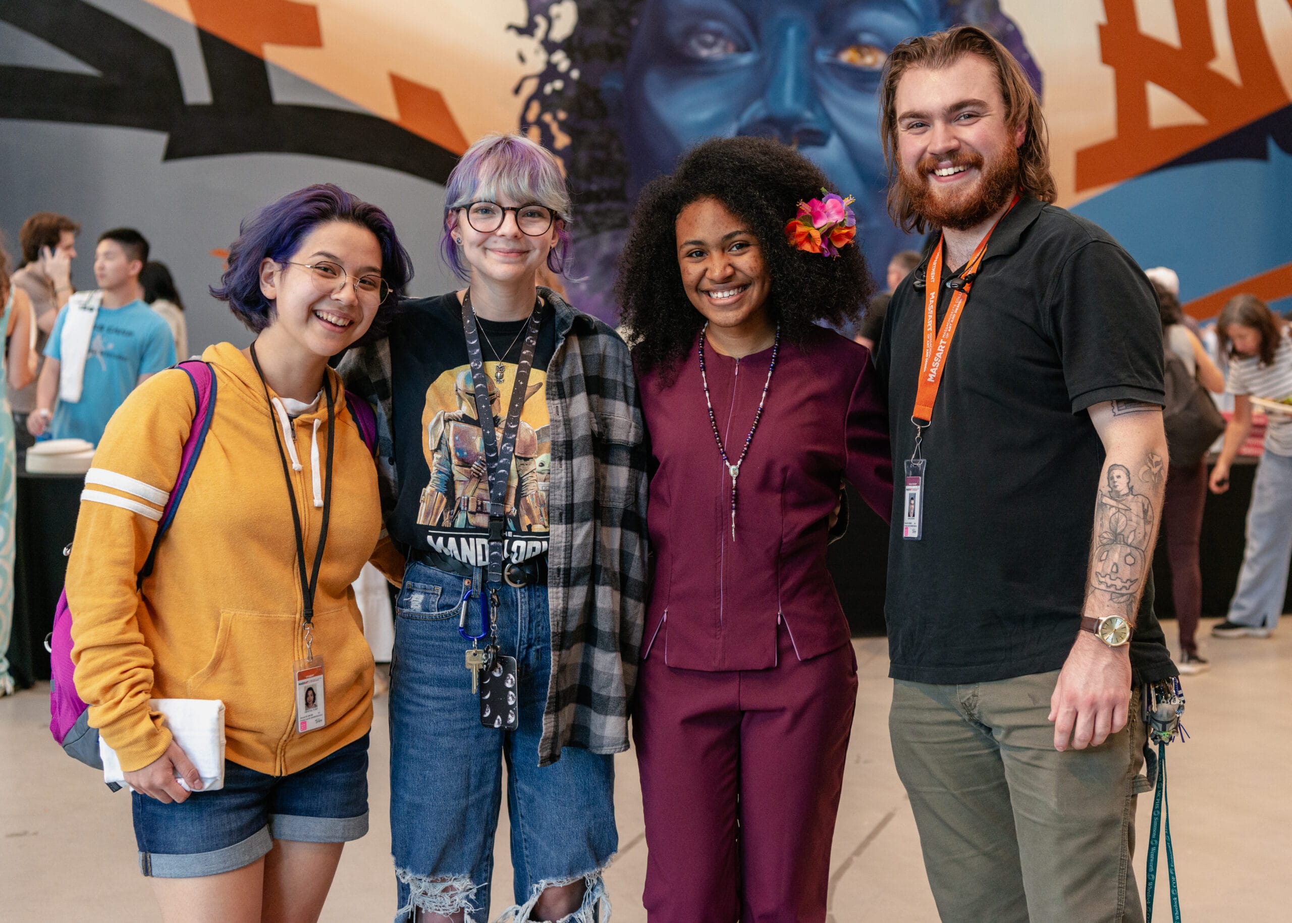 Four people smiling and standing together indoors in a casual setting, capturing the essence of student life. The background features a colorful mural, adding vibrancy to their relaxed and happy demeanor.