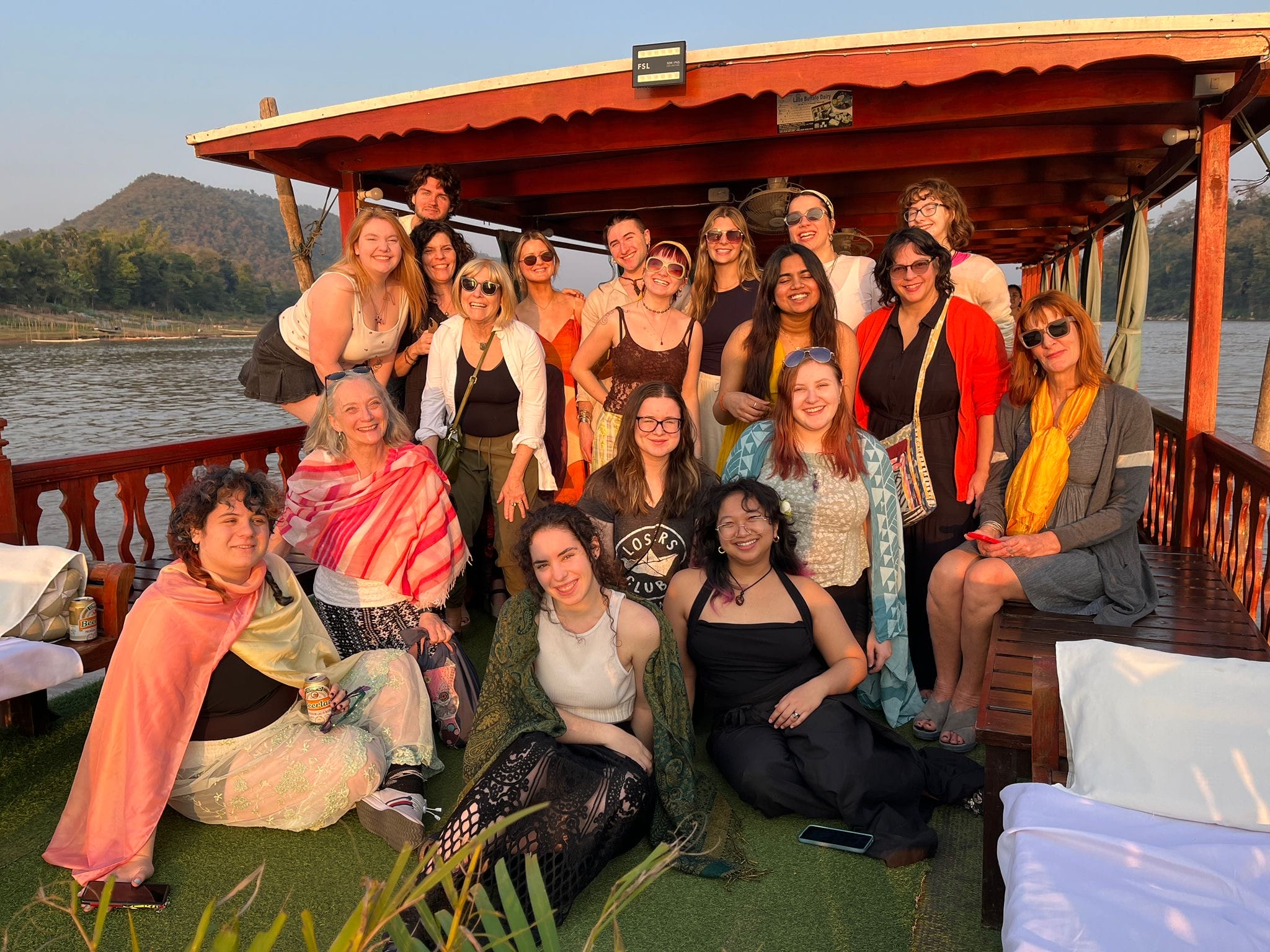 A group of smiling fashion students and professors wearing warm weather clothing gathered on a boat in Laos at the golden hour.