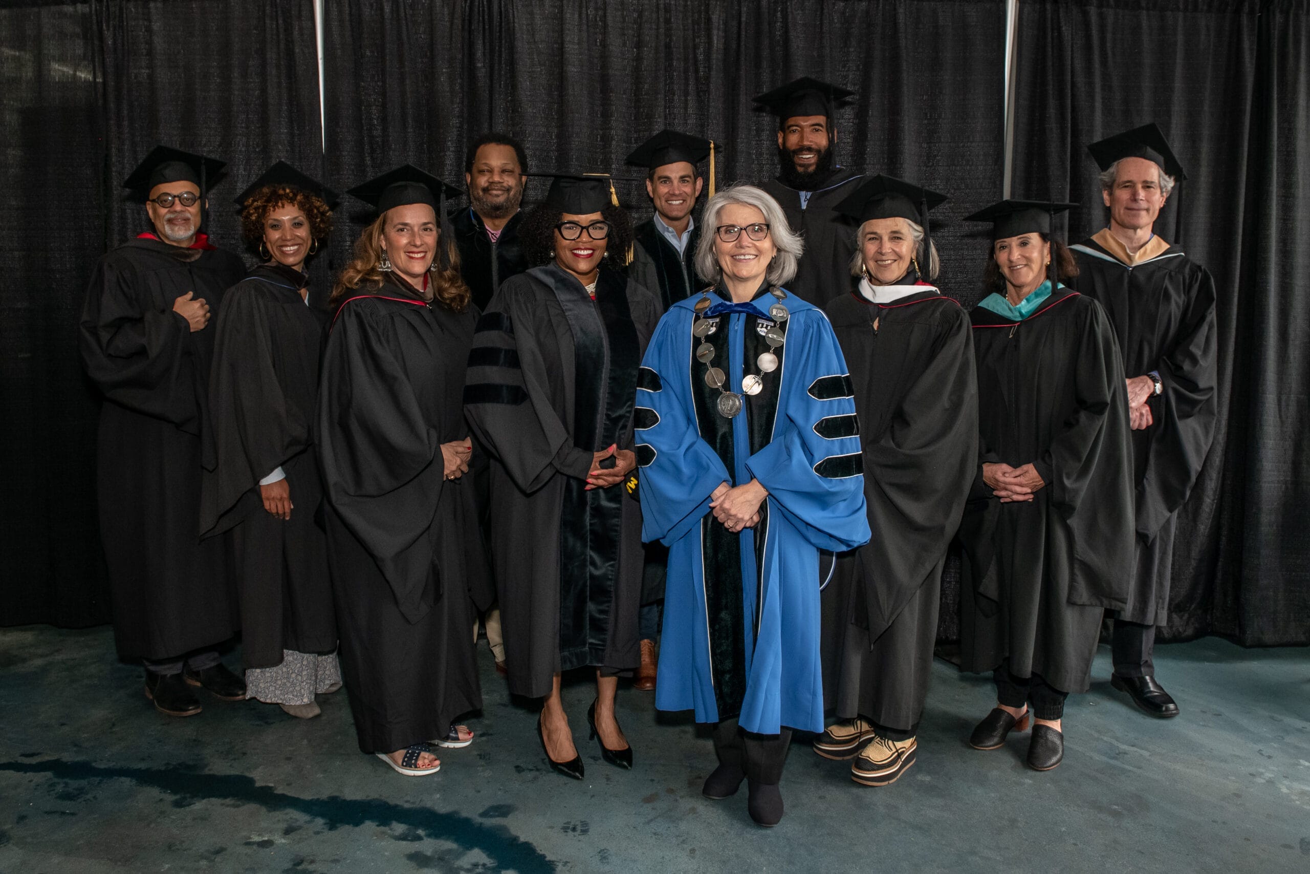 A group of eleven people in graduation robes stands together in front of a black curtain. One person in a light blue robe stands in front, smiling. The rest wear black robes. They are posing for a group photo.