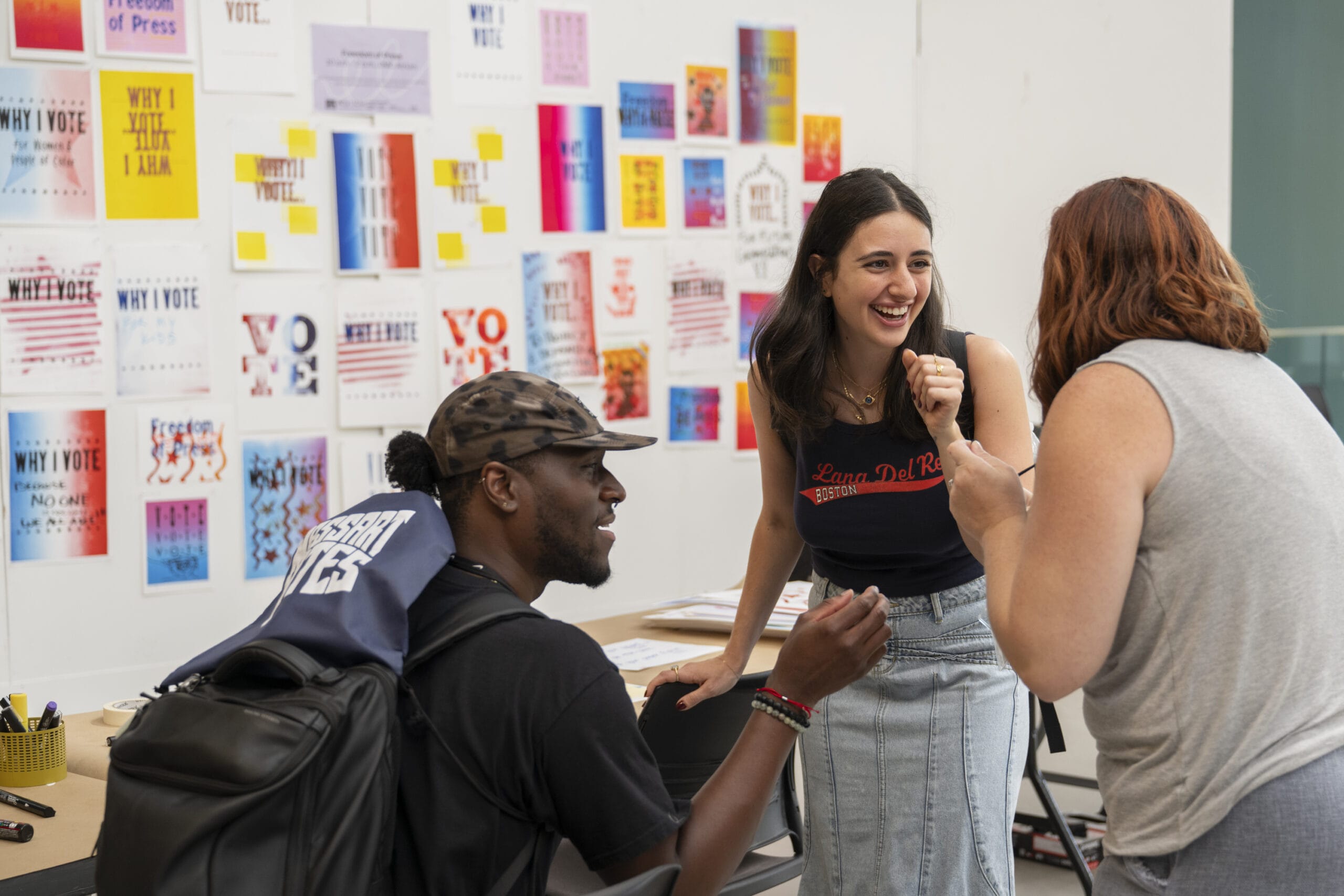 Three people talking and smiling in front of a wall of screen-printed posters that say vote.