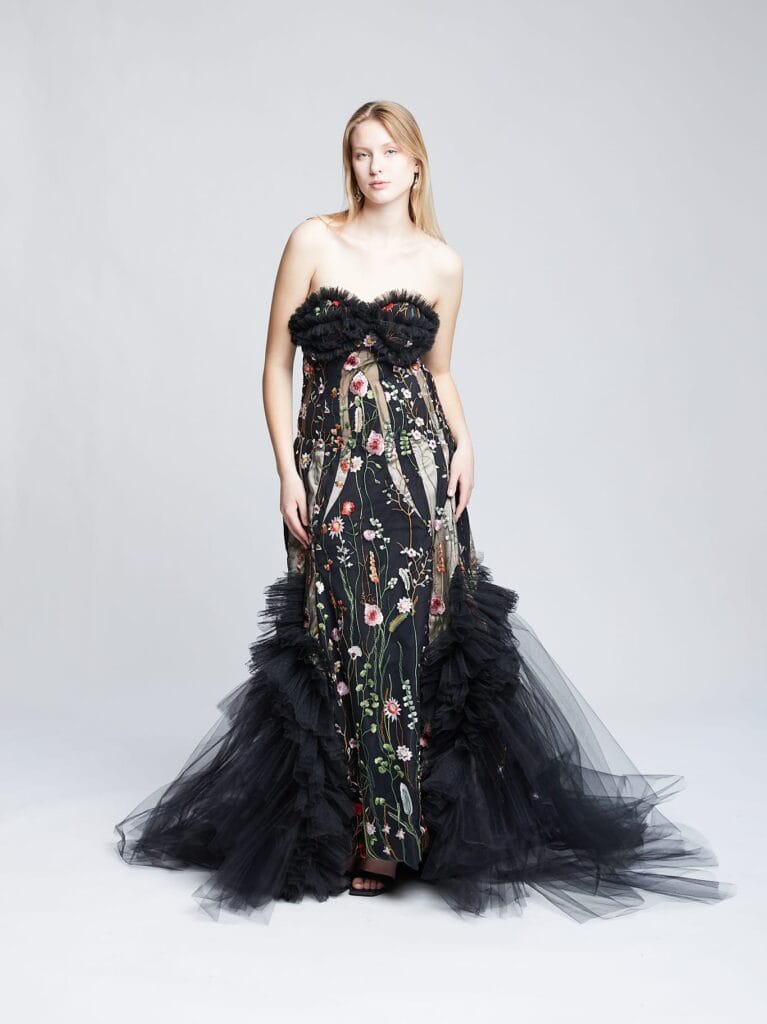 A woman is standing in an elegant, strapless black gown adorned with floral embroidery and tulle details. She is posing against a neutral background. Her expression is serene, and her hair is long and down.