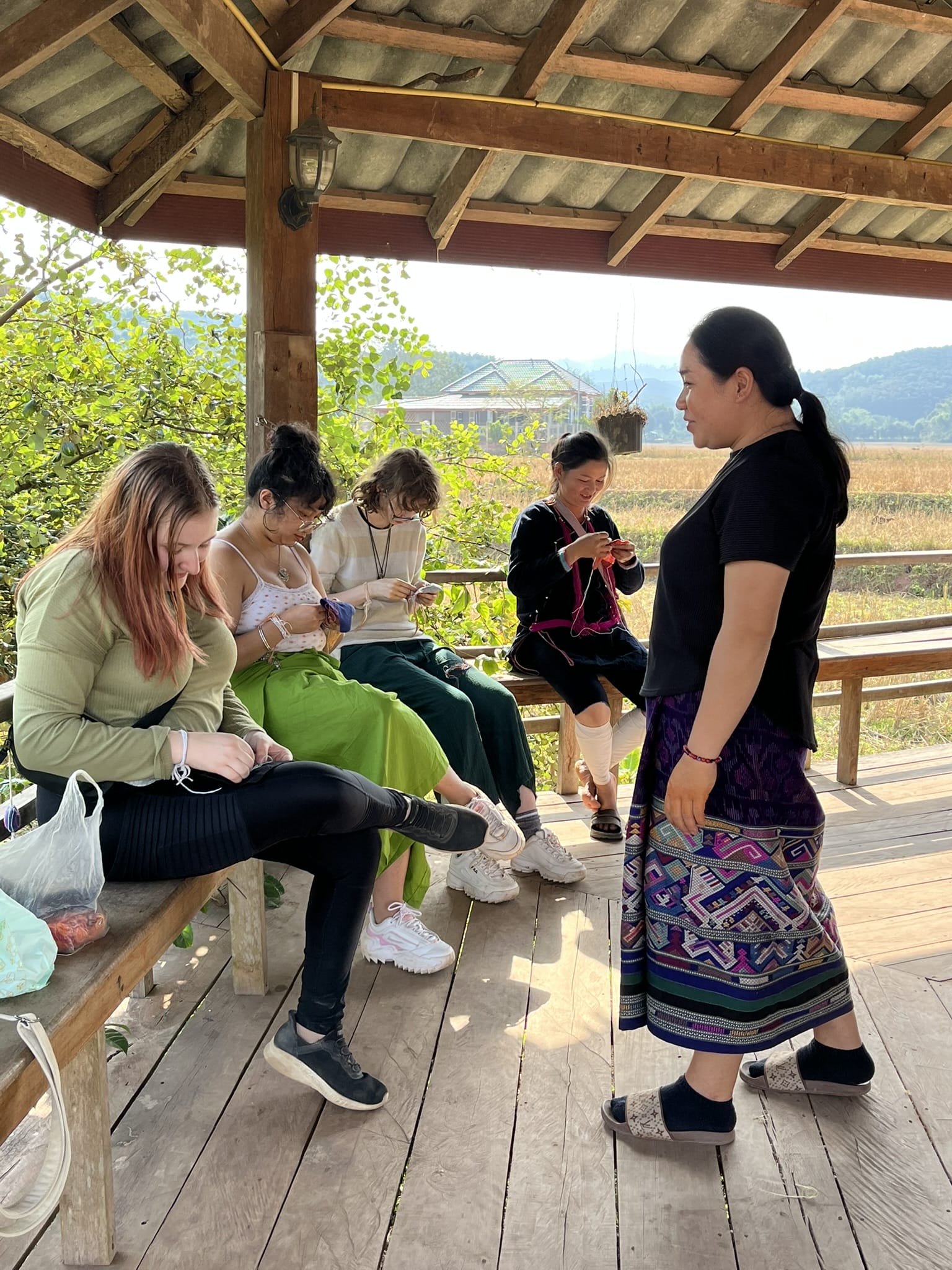 Four people sit on a bench in a covered open-air structure working on embroidery while another woman stands supervising their work.