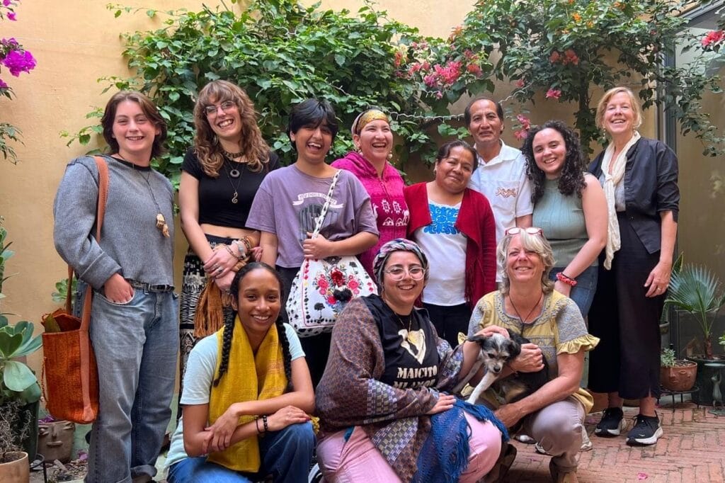 A group of people smiling and standing together in a courtyard with lush plants on the walls, enjoying a sunny day. One person holds a dog. Their camaraderie and shared passion for sustainability are evident in their expressions, as if just returning from inspiring student travel courses.
