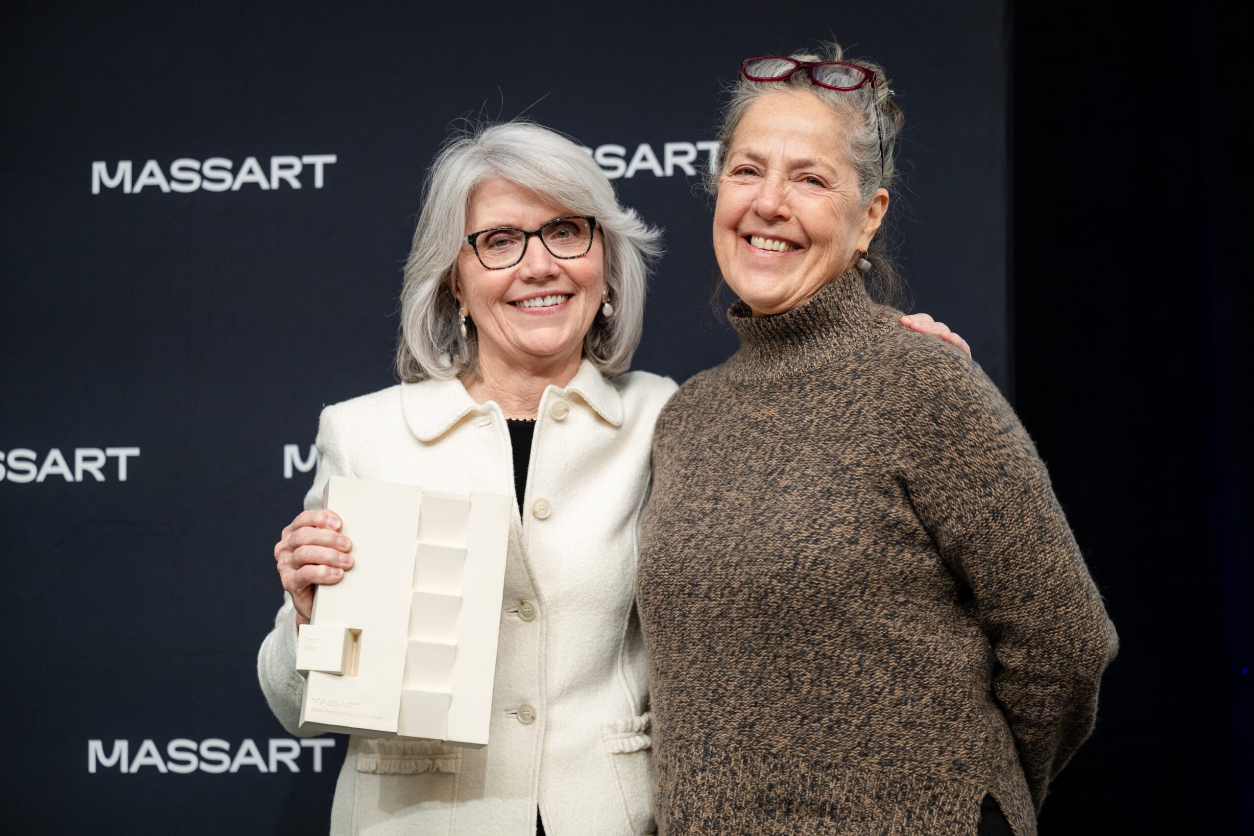 Two women pose smiling with a ceramic award in one of their hands in front of a backdrop that says MassArt.