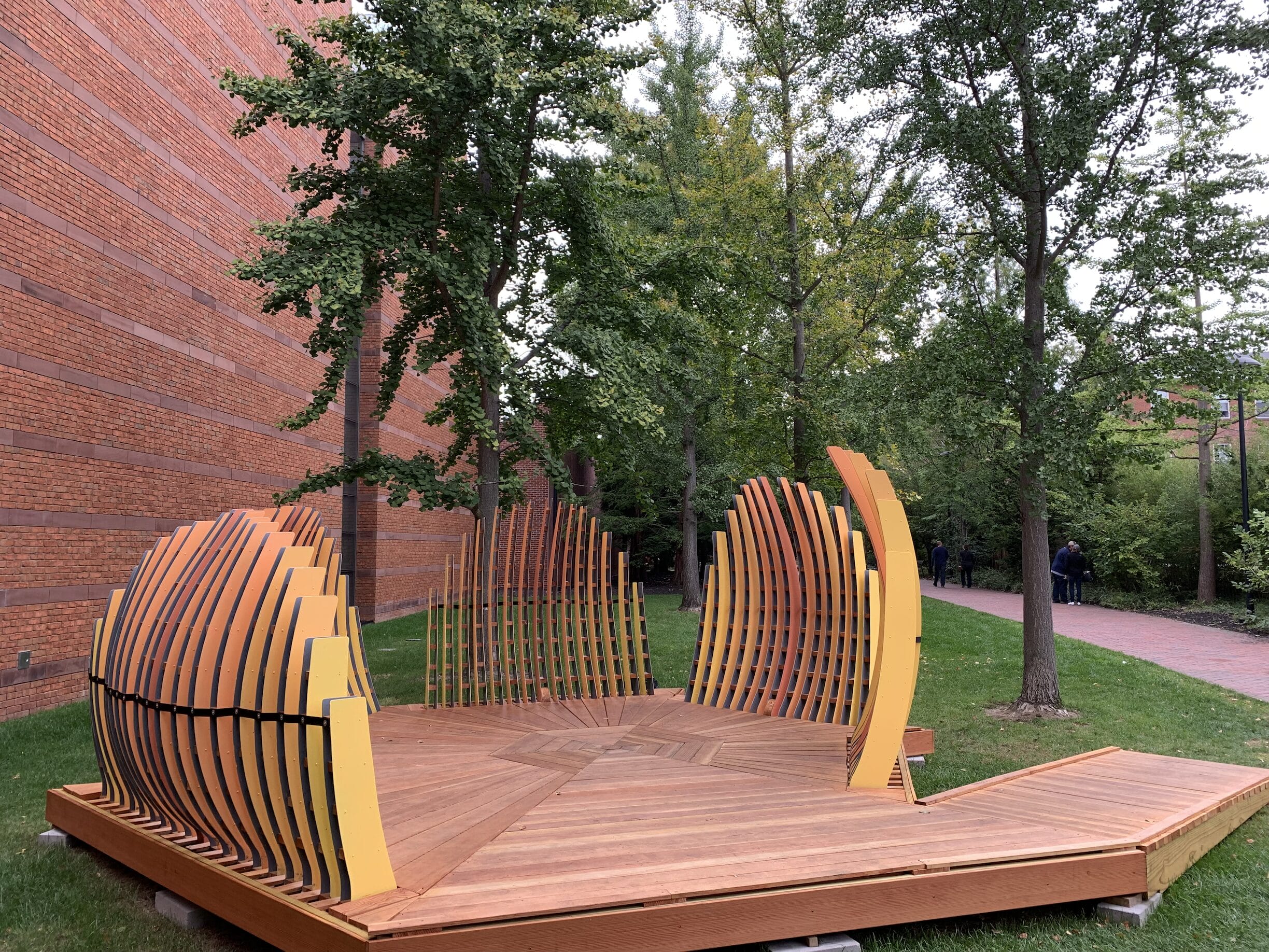 A pavilion structure made of painted steel and wood pictured next to a brick building on the grass, surrounded by trees.