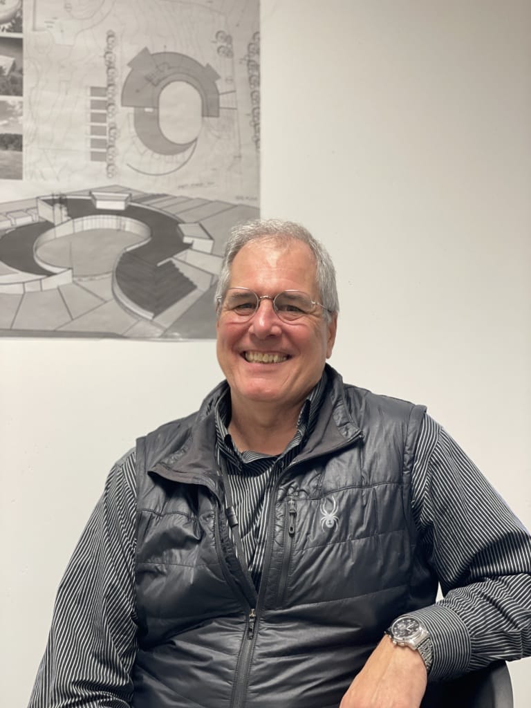 A man with gray hair and silver framed glasses smiles in front of a gray and white architectural artwork on the wall.