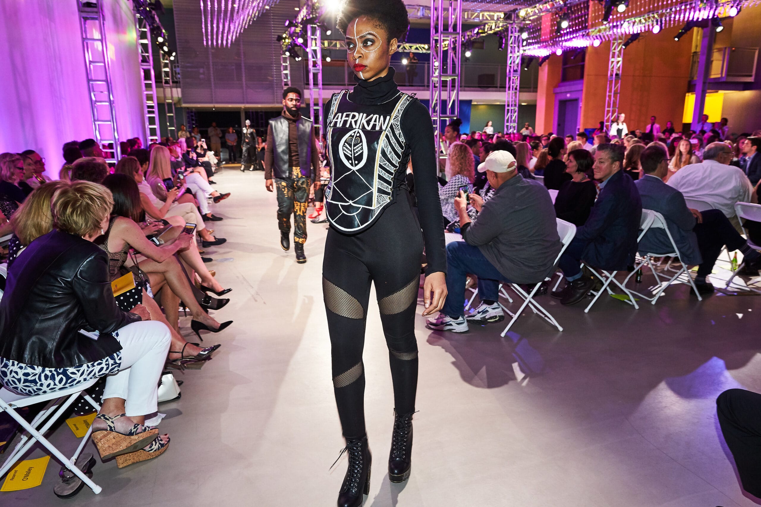 A model wearing a black designed garment with the word "Afrikan" on the front of her chest walks down a runway in a purple lit Design and Media Center at MassArt