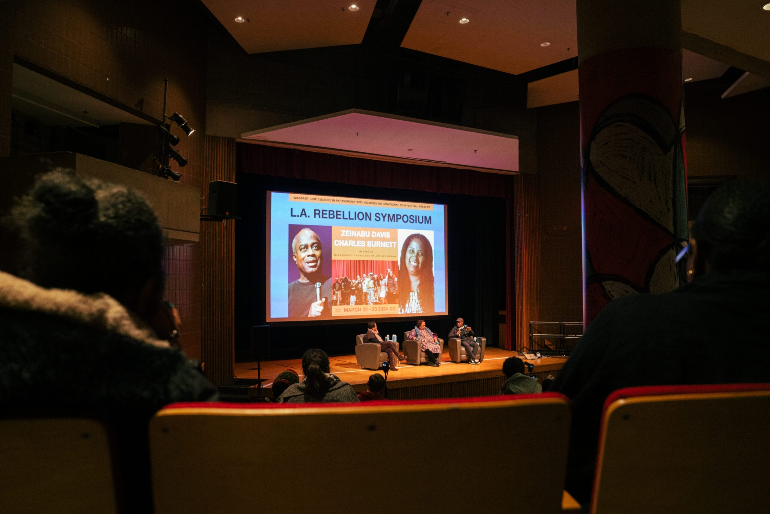 A symposium in an auditorium features a large screen displaying L.A. Rebellion Symposium with images of two visiting artists. Three people are seated onstage, engaged in conversation, offering insightful lectures. The audience watches intently from their seats.