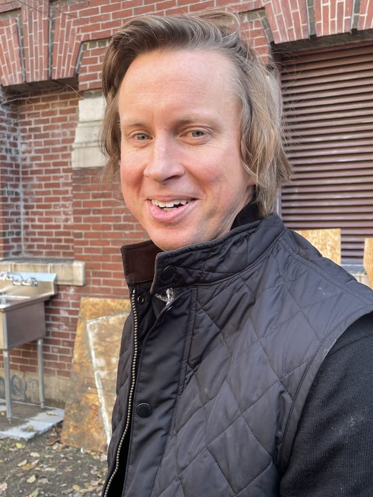 Man in a black quilted vest, outdoors, stands smiling in front of a brick wall and metal sink.