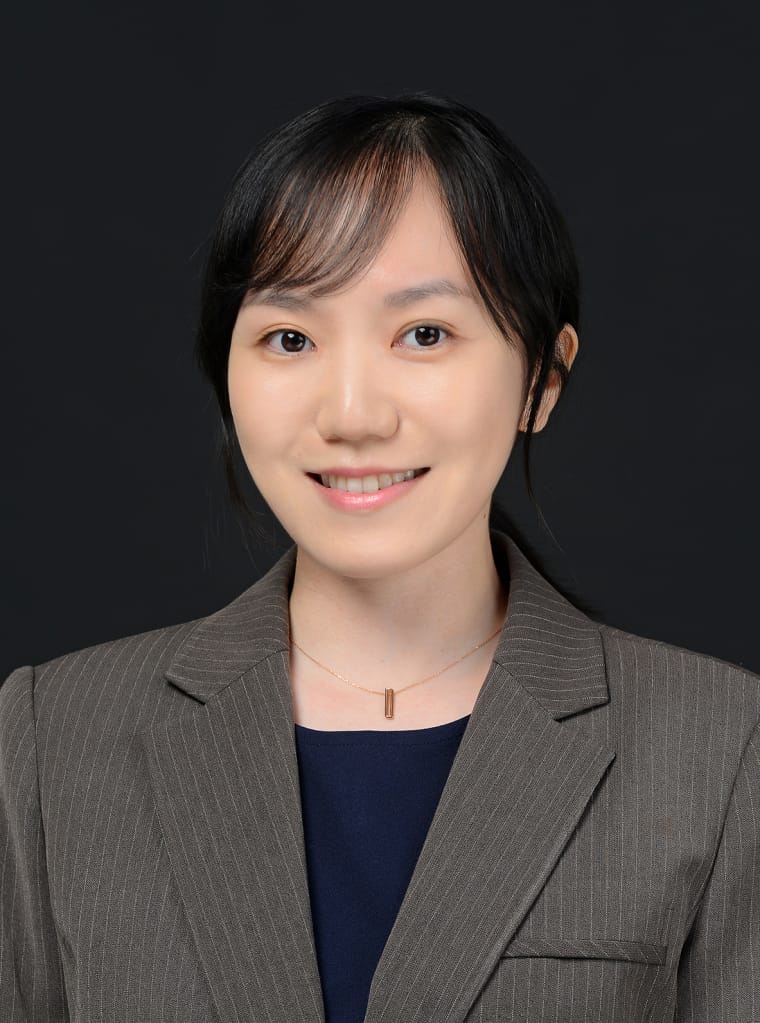 A person with long black hair wearing a pinstripe suit jacket over a dark top smiles against a dark background.