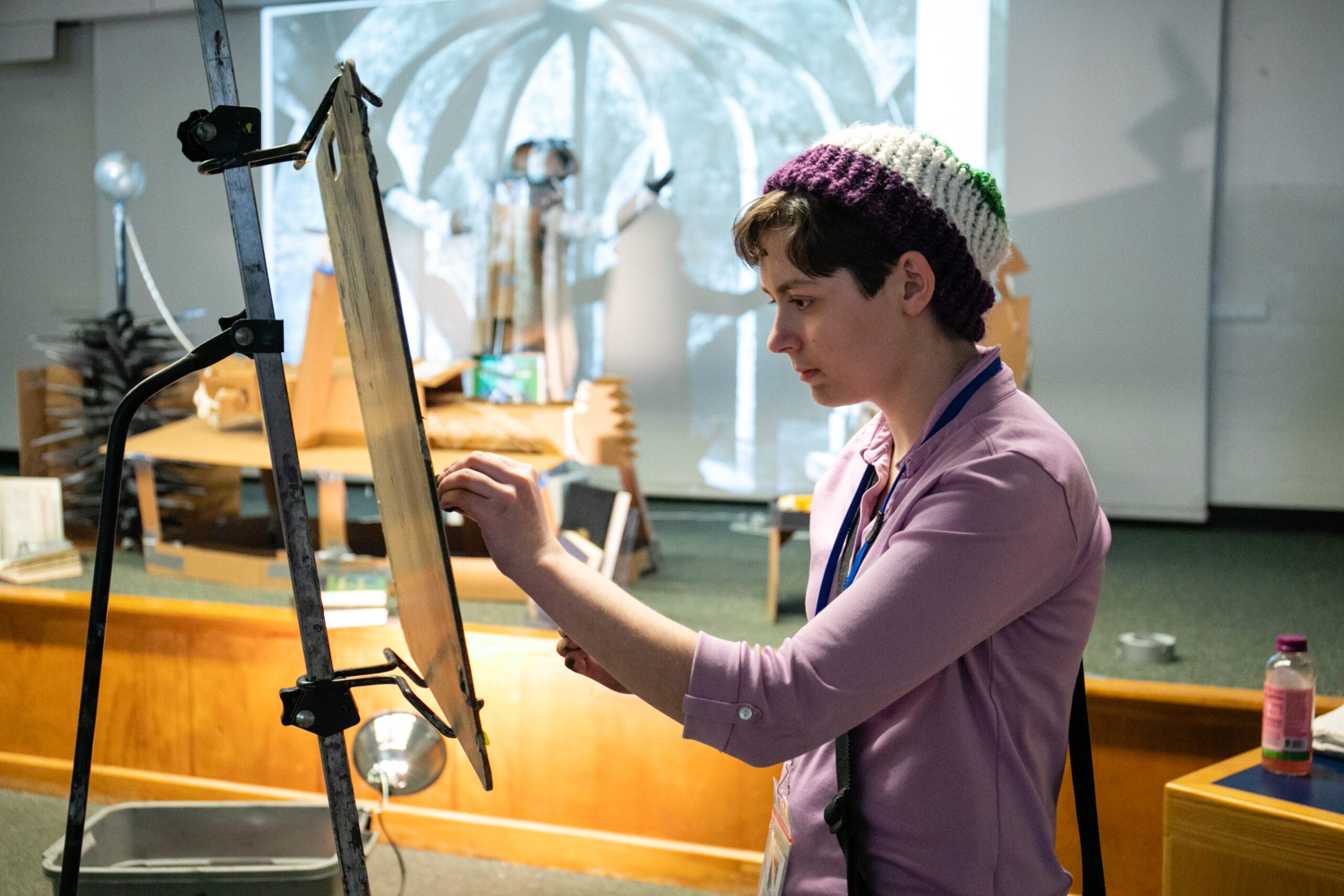 A student wearing a purple shirt and a wool hat stands drawing at an easel.