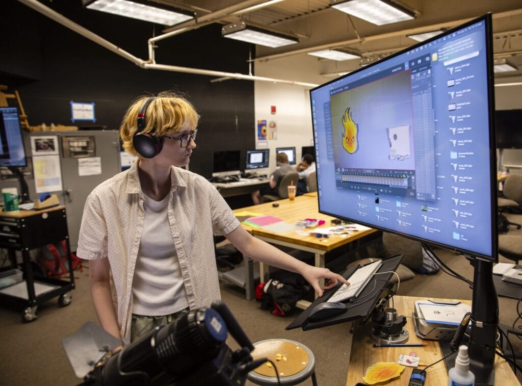 In a studio buzzing with creative energy, a person wearing headphones works on digital art, pointing at a large monitor displaying animation software. This vibrant atmosphere is part of the Summer Intensives, where others also immerse themselves in artistic pursuits at their desks.