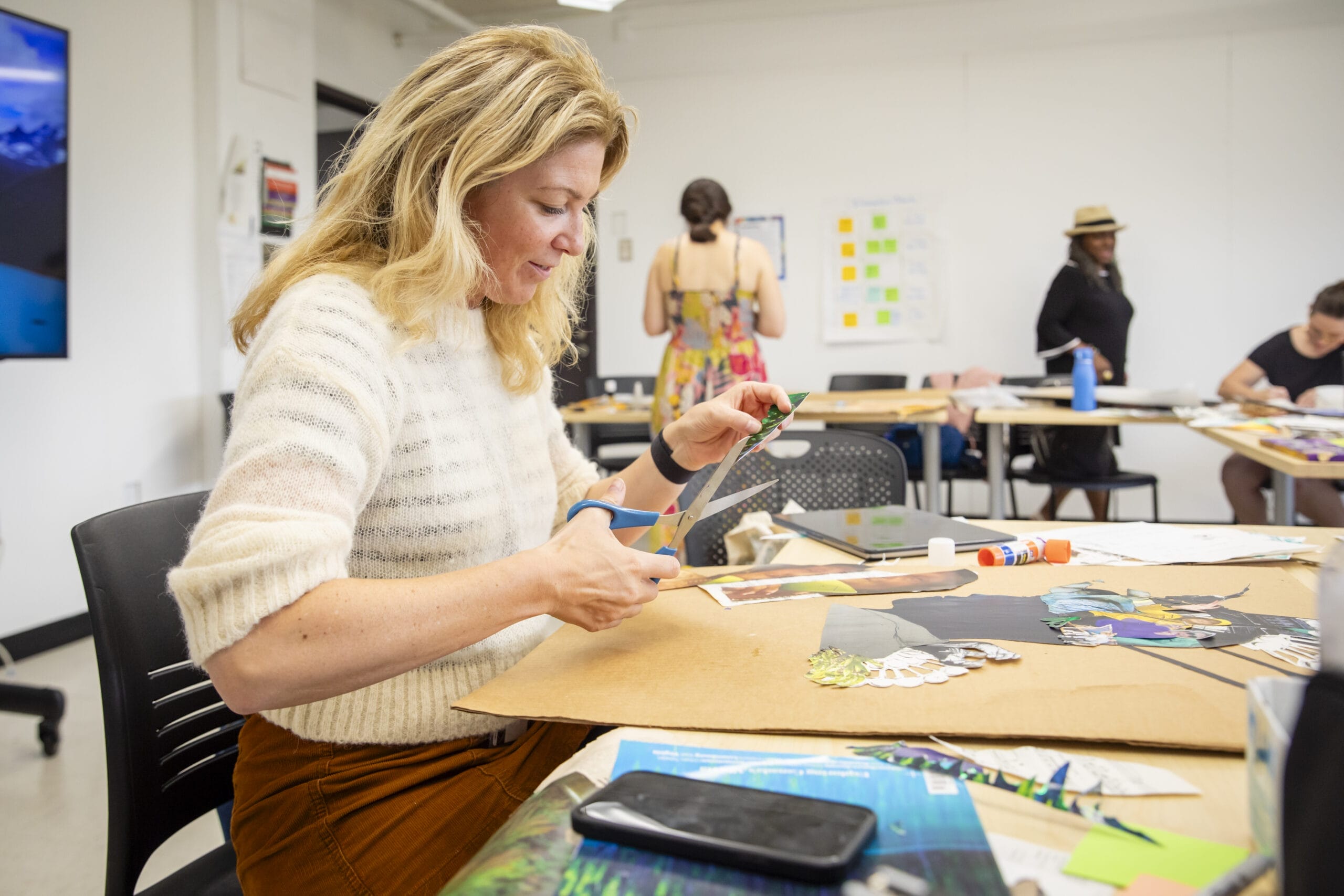 A blond woman wearing a white sweater sits at a table holding scissors and cutting into paper to make a collage.