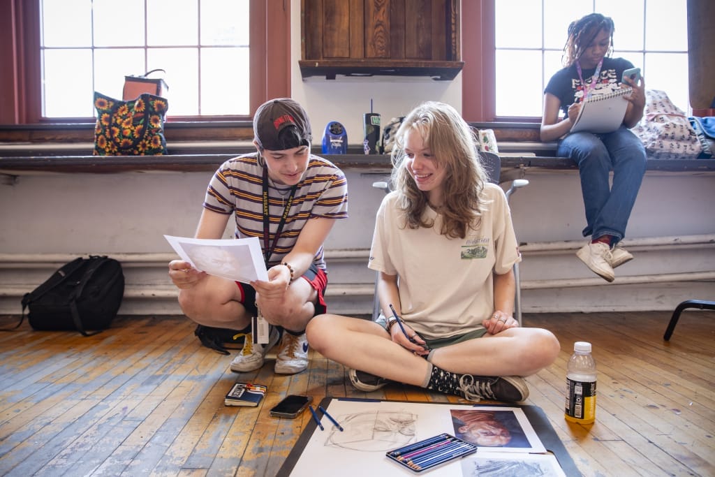 Two young people, one in a striped shirt and cap, the other in a white t-shirt, sit on the studio floor engrossed in drawing. Art supplies are scattered around. During this pre-college summer session, a third person sits on a windowsill scribbling notes.