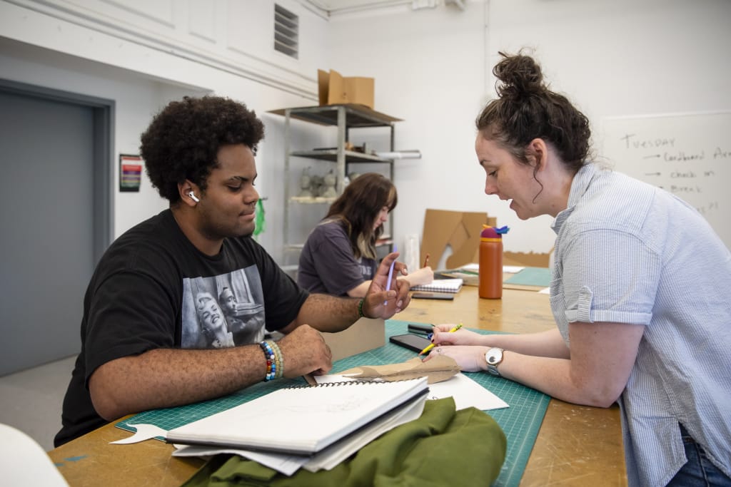 Two people are collaborating at a table brimming with art supplies, including sketchbooks and fabric. One sits, earbuds in, pencil poised; the other stands, gesturing animatedly. In the background of this vibrant Pre-College Summer Studios scene, another person focuses intently on their work.