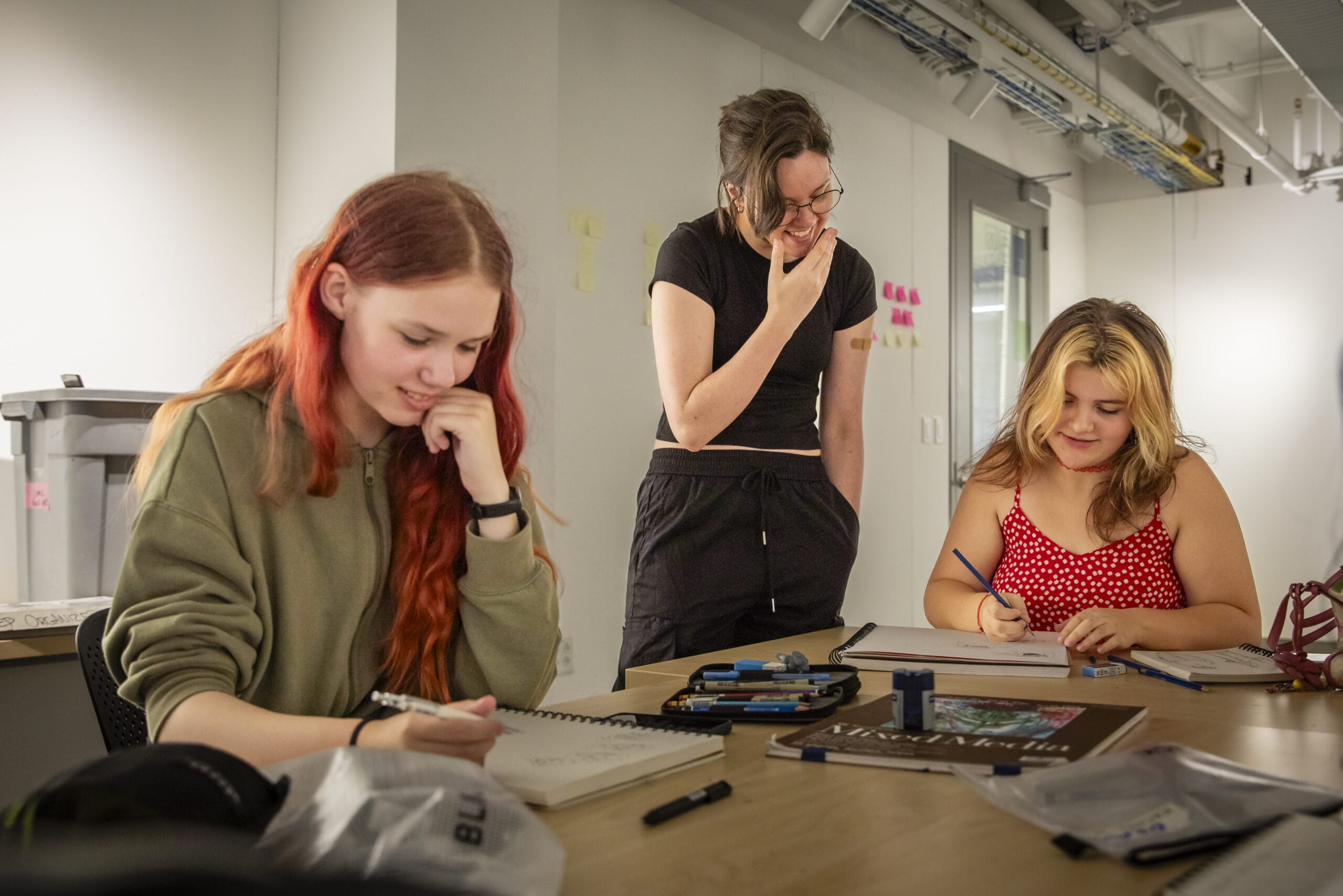 Two girls sit at a desk sketching in their sketchbooks while another woman stands over them giving instructions.