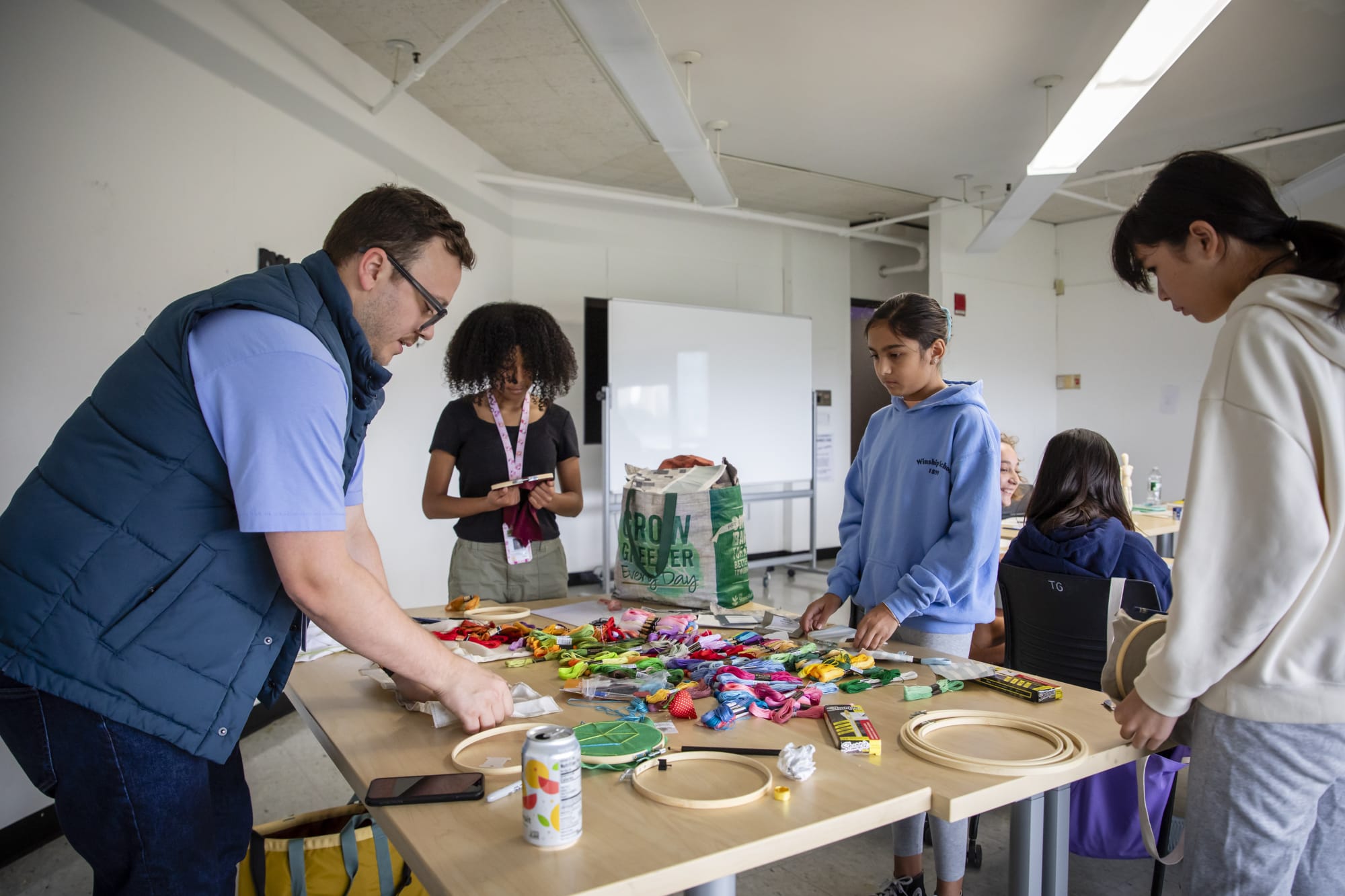 A man with classes hovers over a table covered in crafts supplies and three students stand around the table observing the materials.