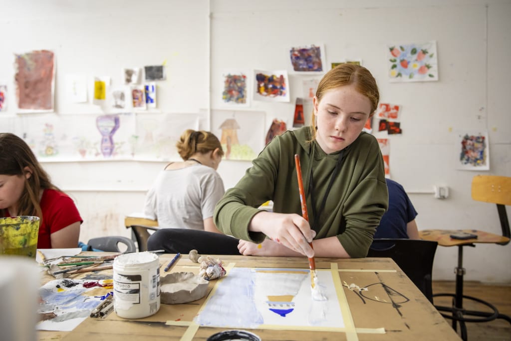 A red-headed student sits at a work table painting in a class.