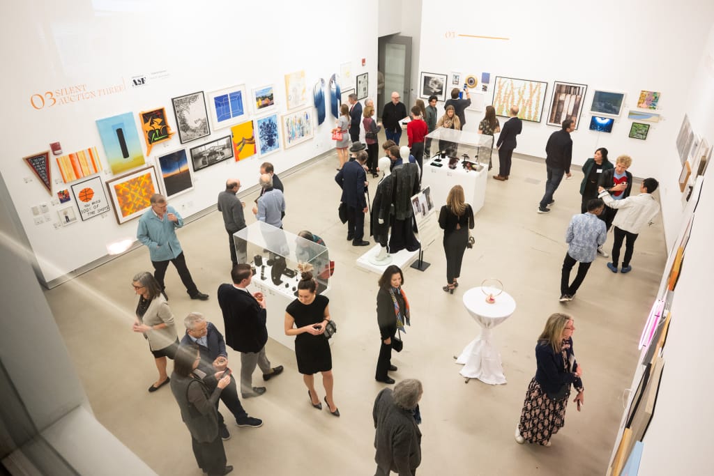 People standing in a white gallery space looking at artwork in the silent auction at MassArt