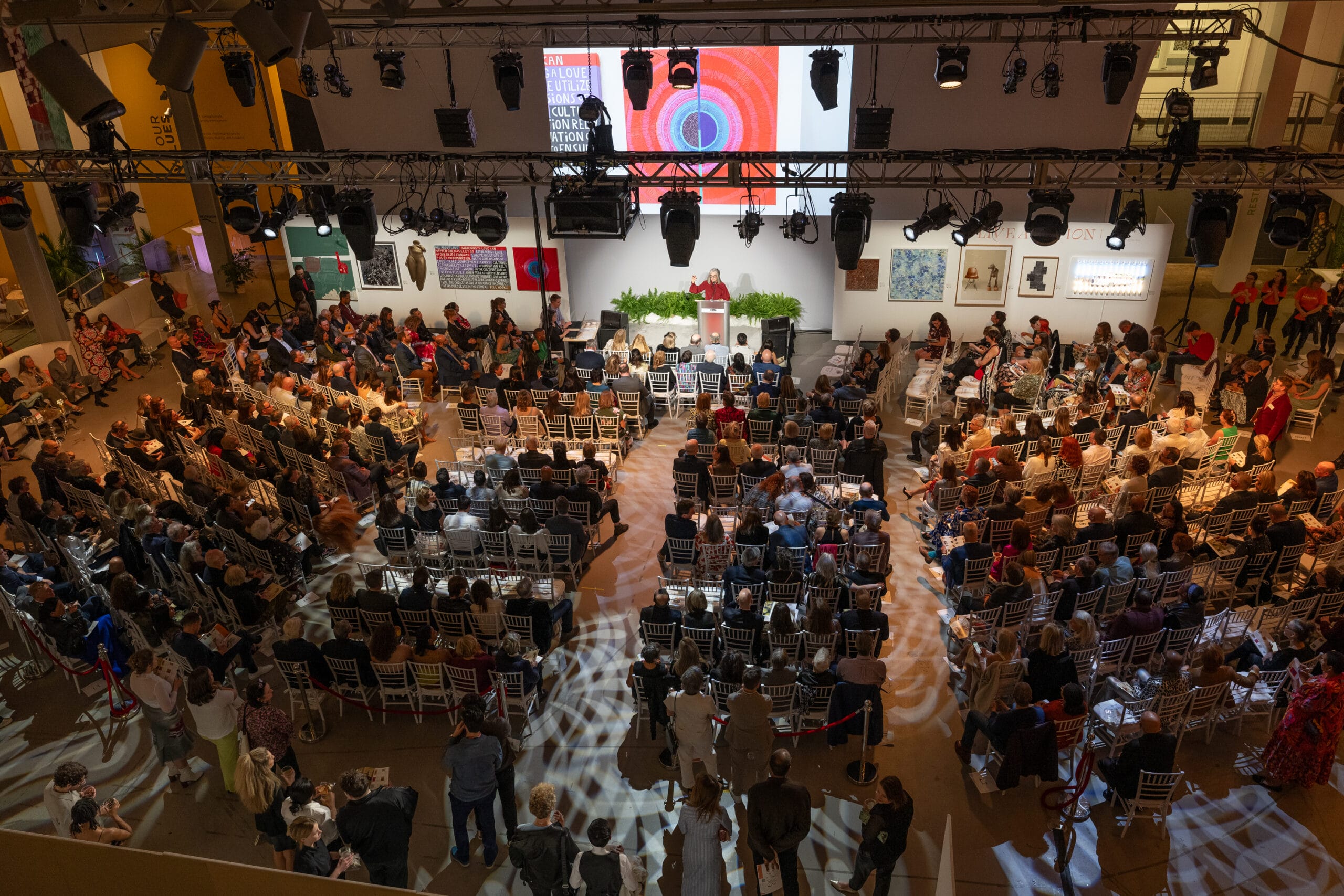 The large indoor event buzzes with anticipation as a full audience faces the stage, where a speaker stands at the podium. The room, well-lit with overhead lights, features a display screen showcasing vibrant red and blue artwork from MassArt. People are engaged and attentive.