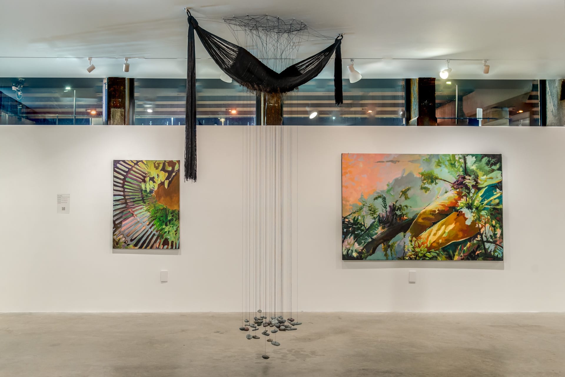 Art gallery with two vibrant nature-themed paintings on a white wall. A black hammock-like installation hangs from the ceiling, with strings extending to scattered stones on the floor below. Bright and serene atmosphere.