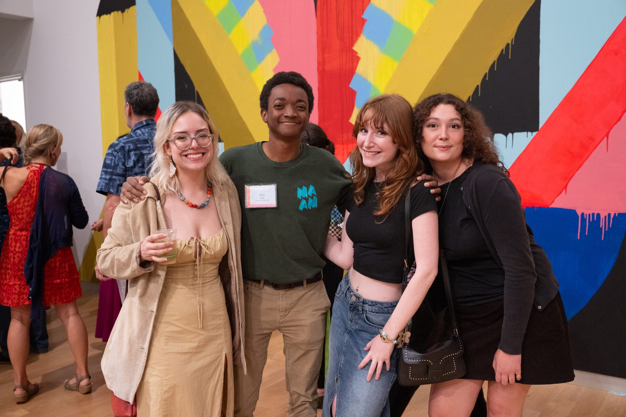 A group of four people poses and smiles in front of a vibrant abstract mural at MAAM, celebrating five years of captivating art. They seem to be enjoying themselves, with one person holding a drink. Other visitors are seen mingling in the background.