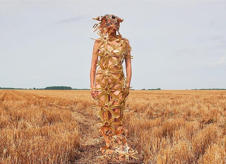 A woman stands in a field of dry grass covered only by woven reeds.