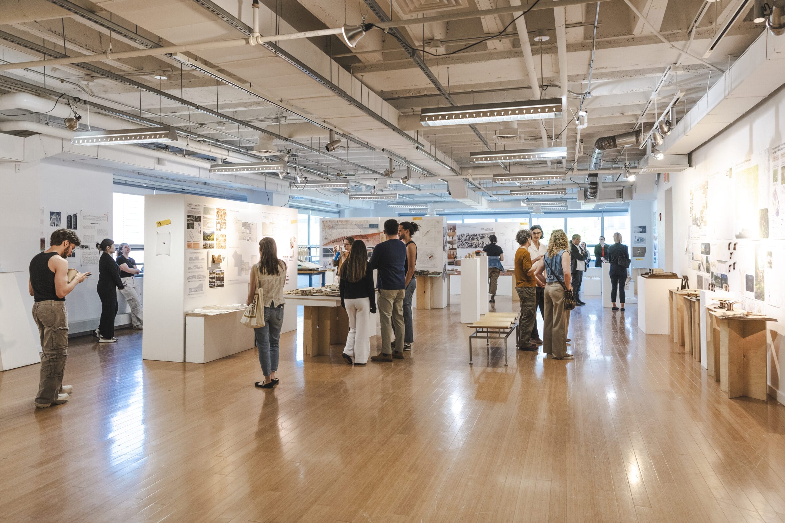 A group of undergraduates are viewing an art exhibition in a spacious gallery. The room has white walls and wooden floors, with displays of various artworks and models. Bright overhead lighting illuminates the space, as visitors are spread throughout the room.