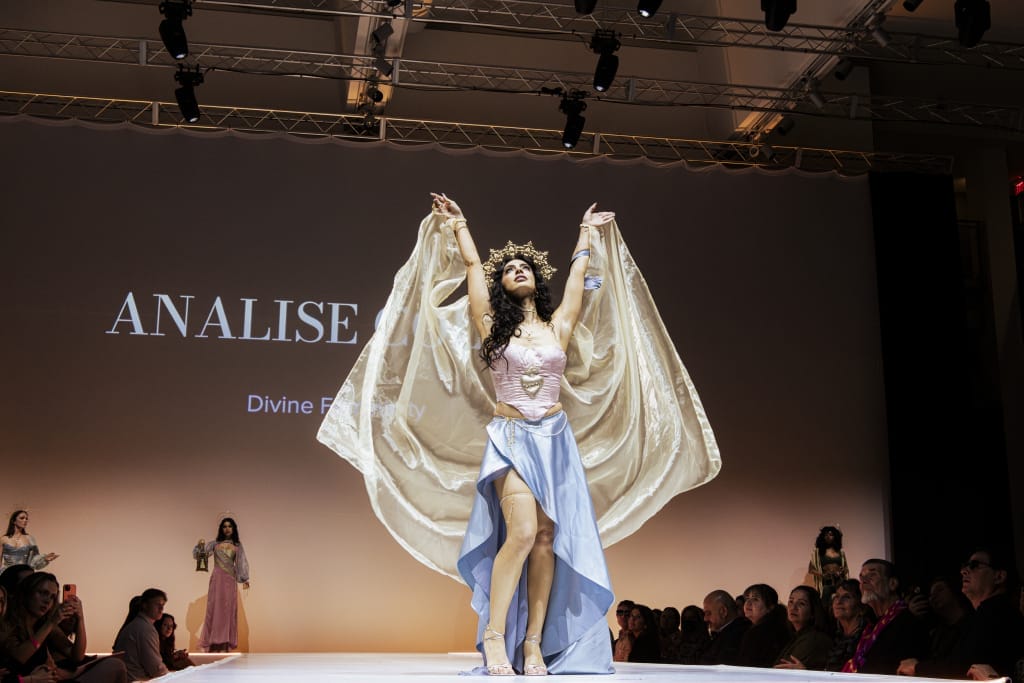 A model on a runway wears a flowing gown with a long cape and a crown. She strikes a dramatic pose with arms raised. The background features the text ANALISE and Divine Femininity. Spectators sit on either side of the runway.